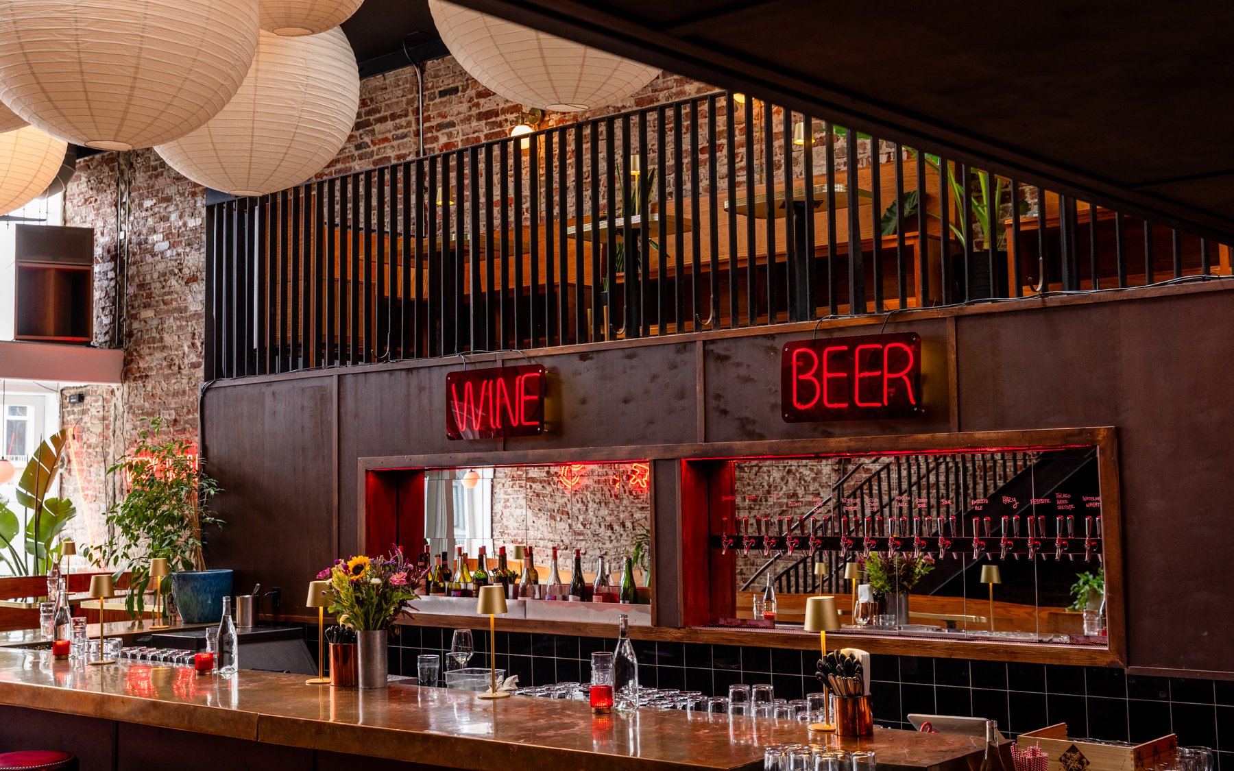 Interior of a bar or restaurant with exposed brick walls, large hanging paper lanterns, and neon signs reading 'WINE' and 'BEER.' There is a bar counter with bottles and glasses, and a second-floor balcony with plants and seating.