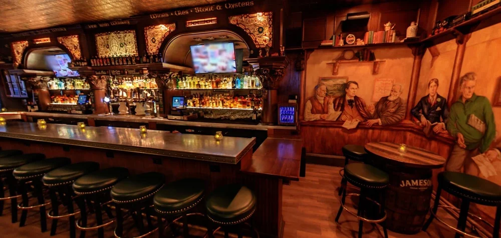 A cozy bar with a long counter and several barstools, a mural of four people talking and sharing stories, shelves of liquor bottles, TV screens, and warm ambient lighting.