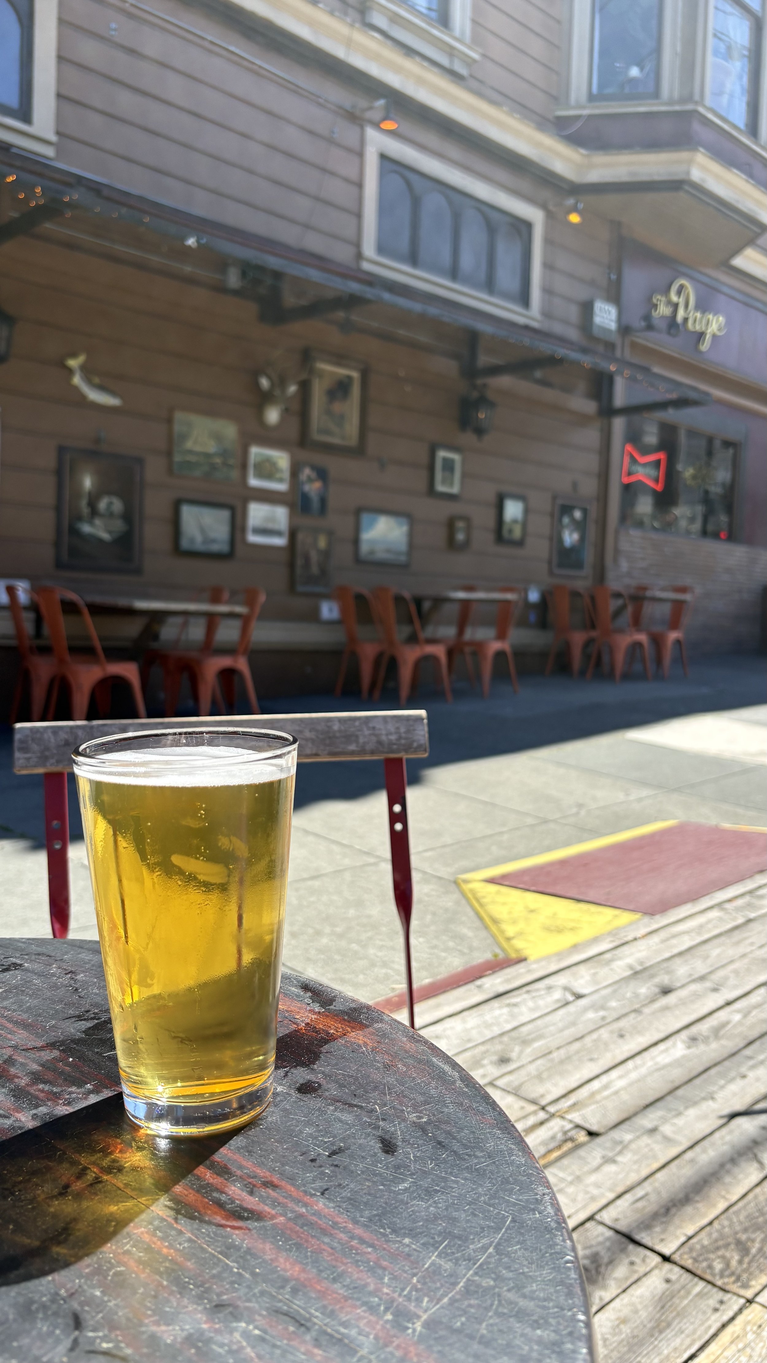 A glass of beer on a small round table outside a restaurant or bar with wooden exterior walls, framed pictures, and signs. There are red chairs and a sidewalk with a yellow and red ramp or step.