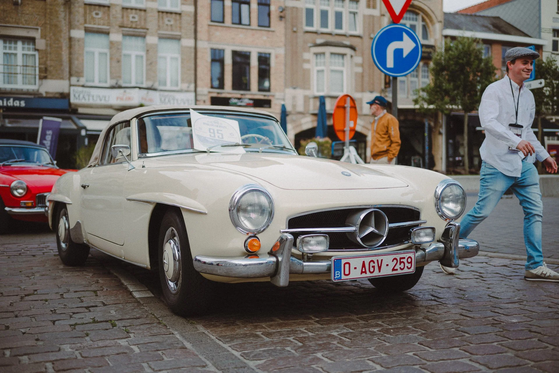 Een klassieke witte sportwagen met Belgisch kenteken, geparkeerd op een stenen straat, met mensen eromheen, in een stedelijke omgeving met historische gebouwen.