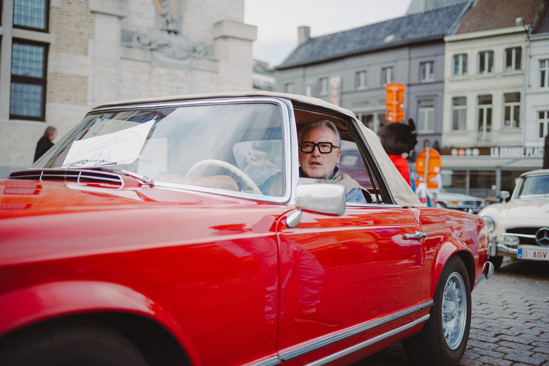 Man met grijs haar en zwarte bril rijdt in een rode klassieke auto op een straat met historische gebouwen op de achtergrond.