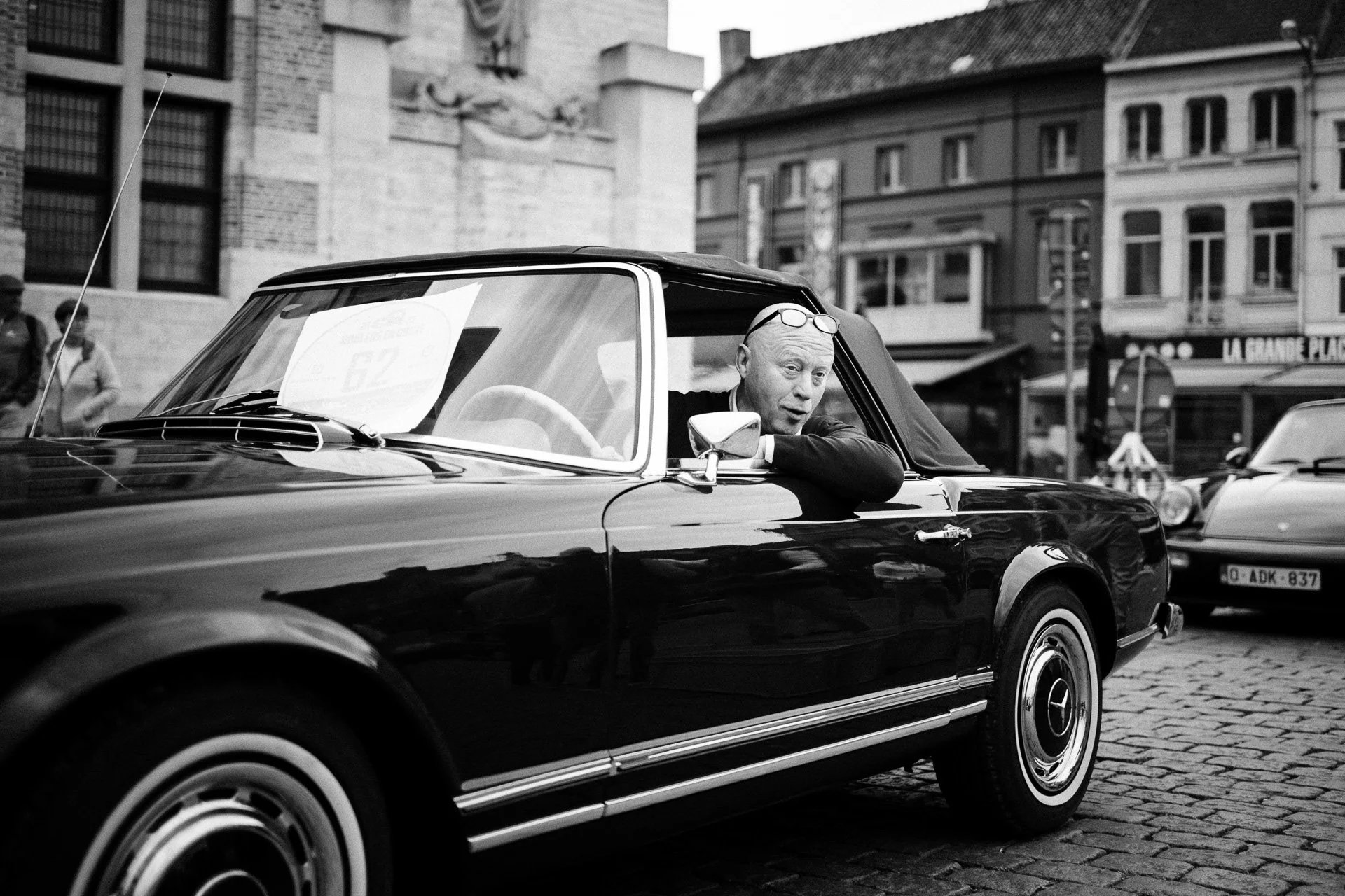 Zwart-witte foto van een man die in een vintage auto zit, met de arm op de portierleuning leunend, in een stadscentrum met historische gebouwen op de achtergrond.