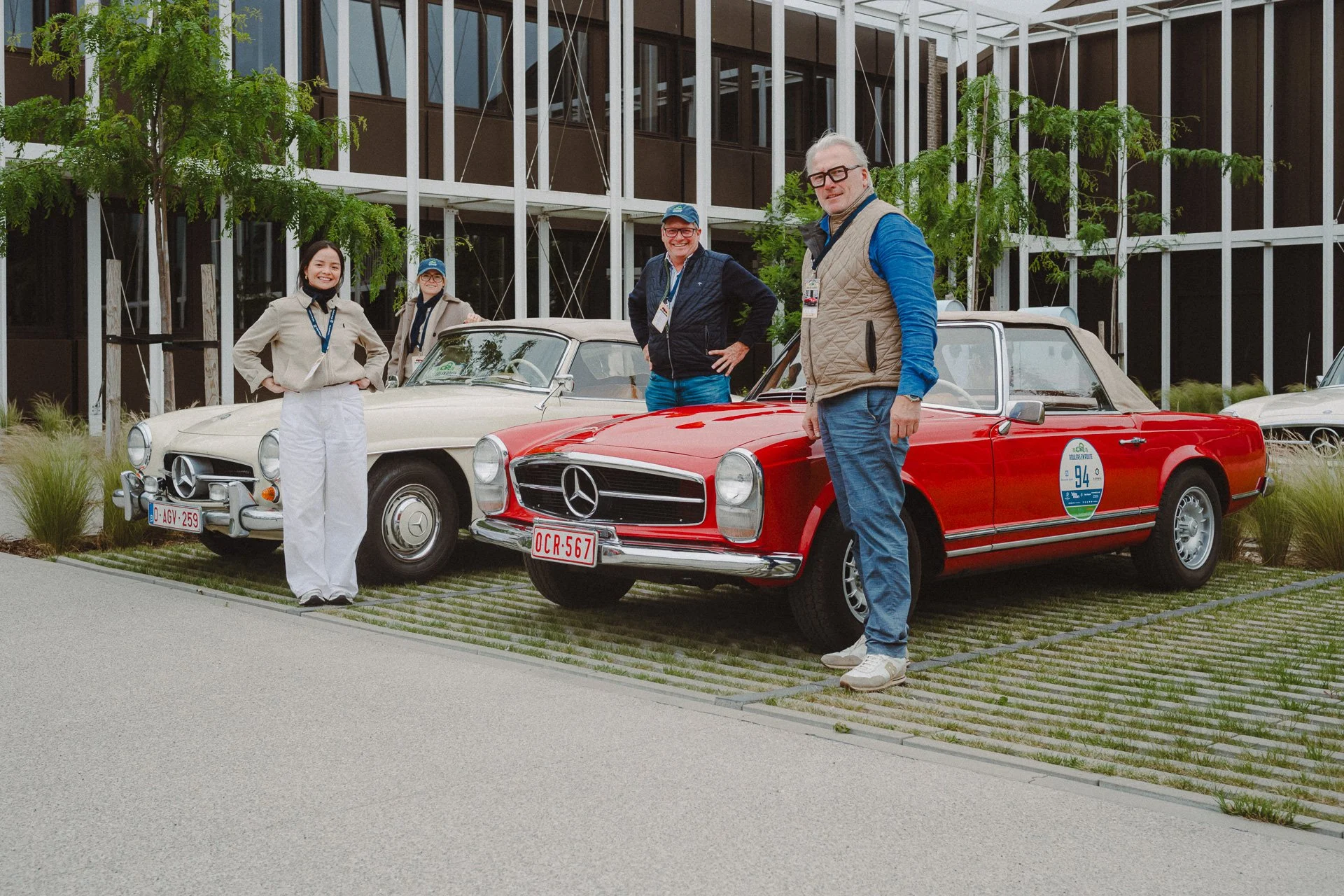 Vier mensen staan naast twee klassieke Mercedes-Benz auto's bij een autoshow of evenement. Ze lachen en poseren voor de camera. De achtergrond toont een modern gebouw met veel glas en groene bomen.