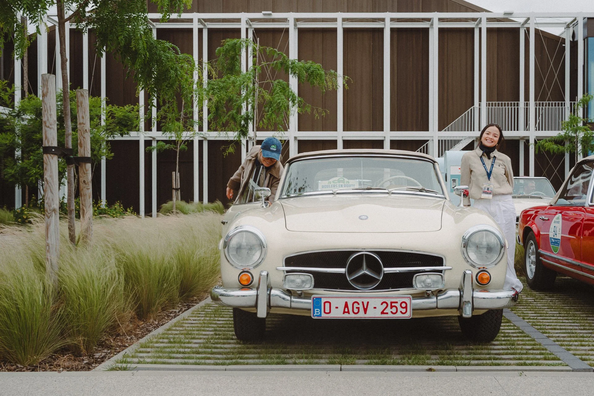 Een wit vintage Mercedes-Benz auto met Kentekenplaat 0-AGV-259, voor een modern gebouw met houten en witte structuur, met twee vrouwen naast auto, één meisje met beige jas en zwart masker, lachend, en een vrouw met blauwe pet die iets in de auto onde