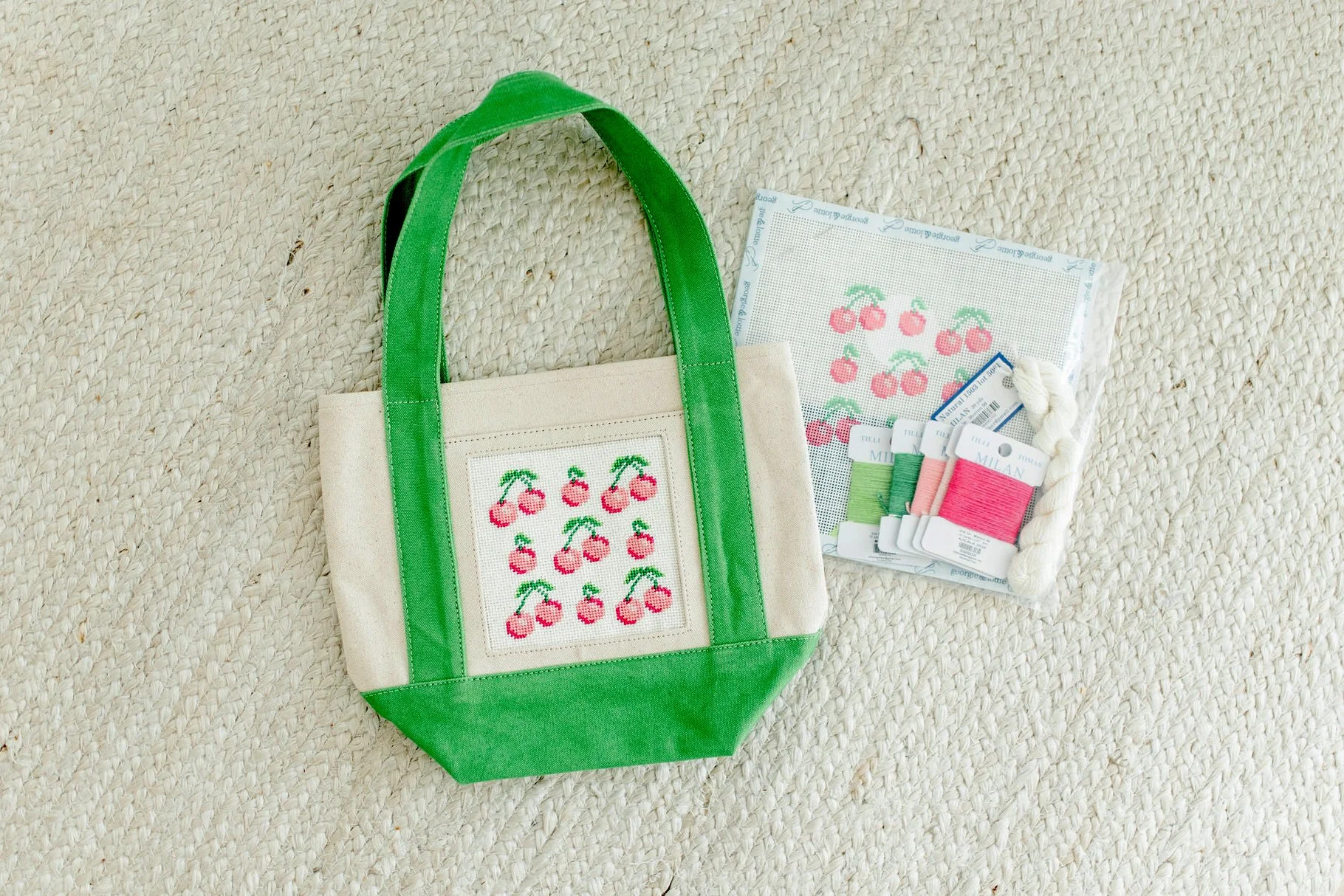 A cream-colored tote bag with green handles and a cross-stitch design of cherries on the front, placed on a textured beige surface. Next to the bag are embroidery floss and printed cross-stitch patterns of cherries.