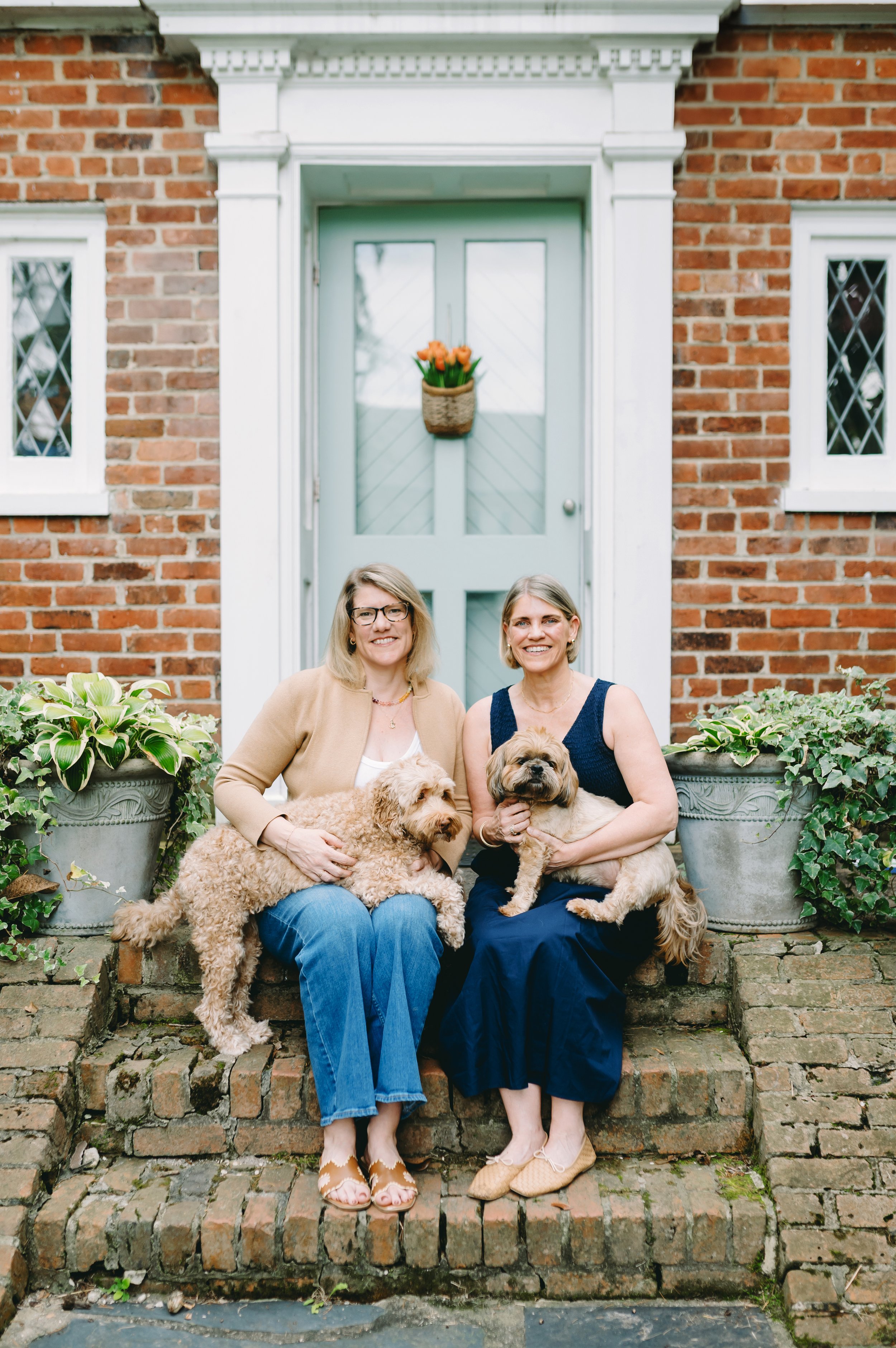 Two women sitting on brick steps in front of a house with brick walls and light blue door, each holding a dog, with potted plants on each side.