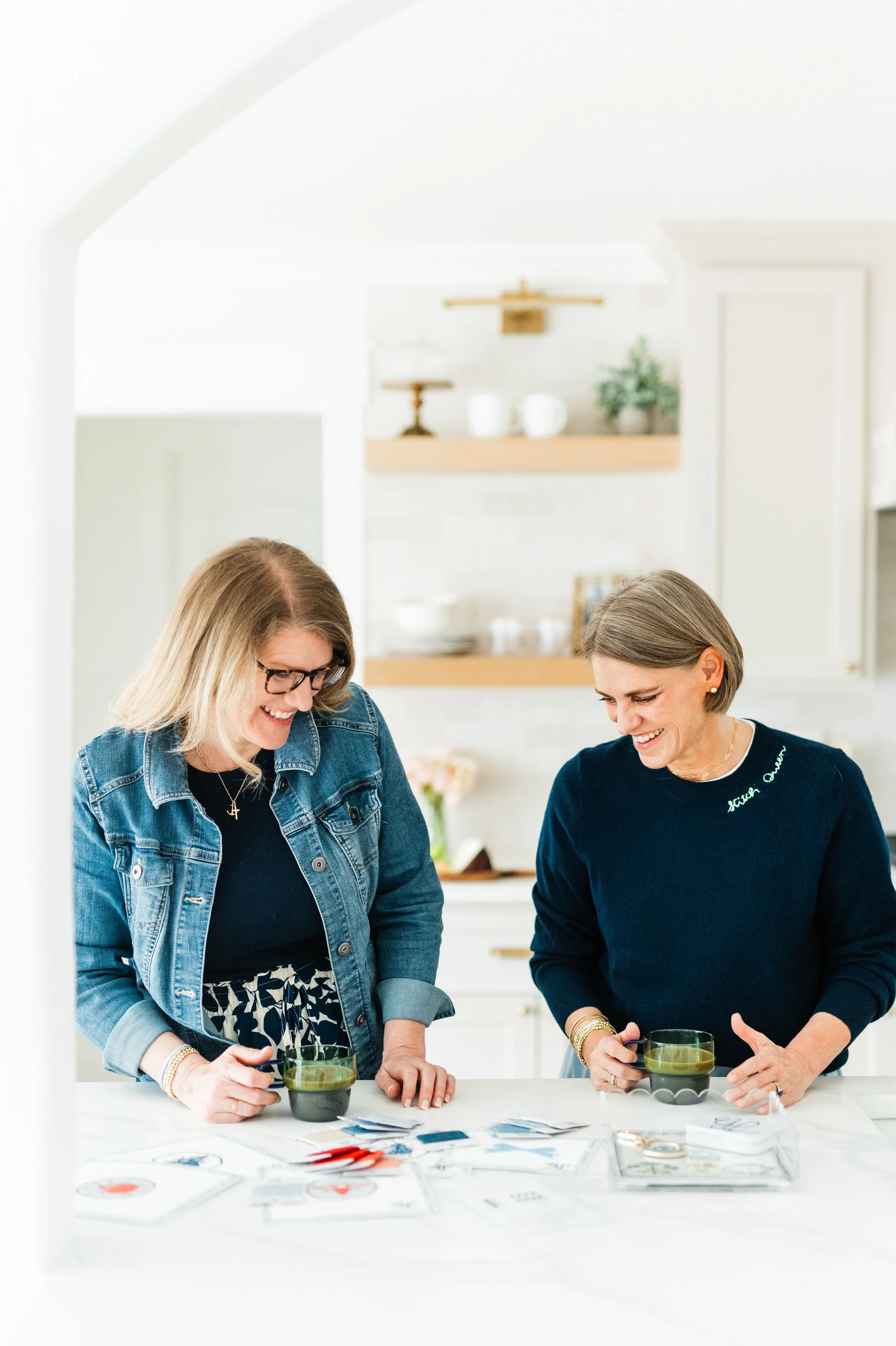 Two women with shoulder-length hair, one with glasses, and the other with jewelry, are standing at a white table with cups and cards, smiling and engaged in an activity in a bright, modern kitchen.