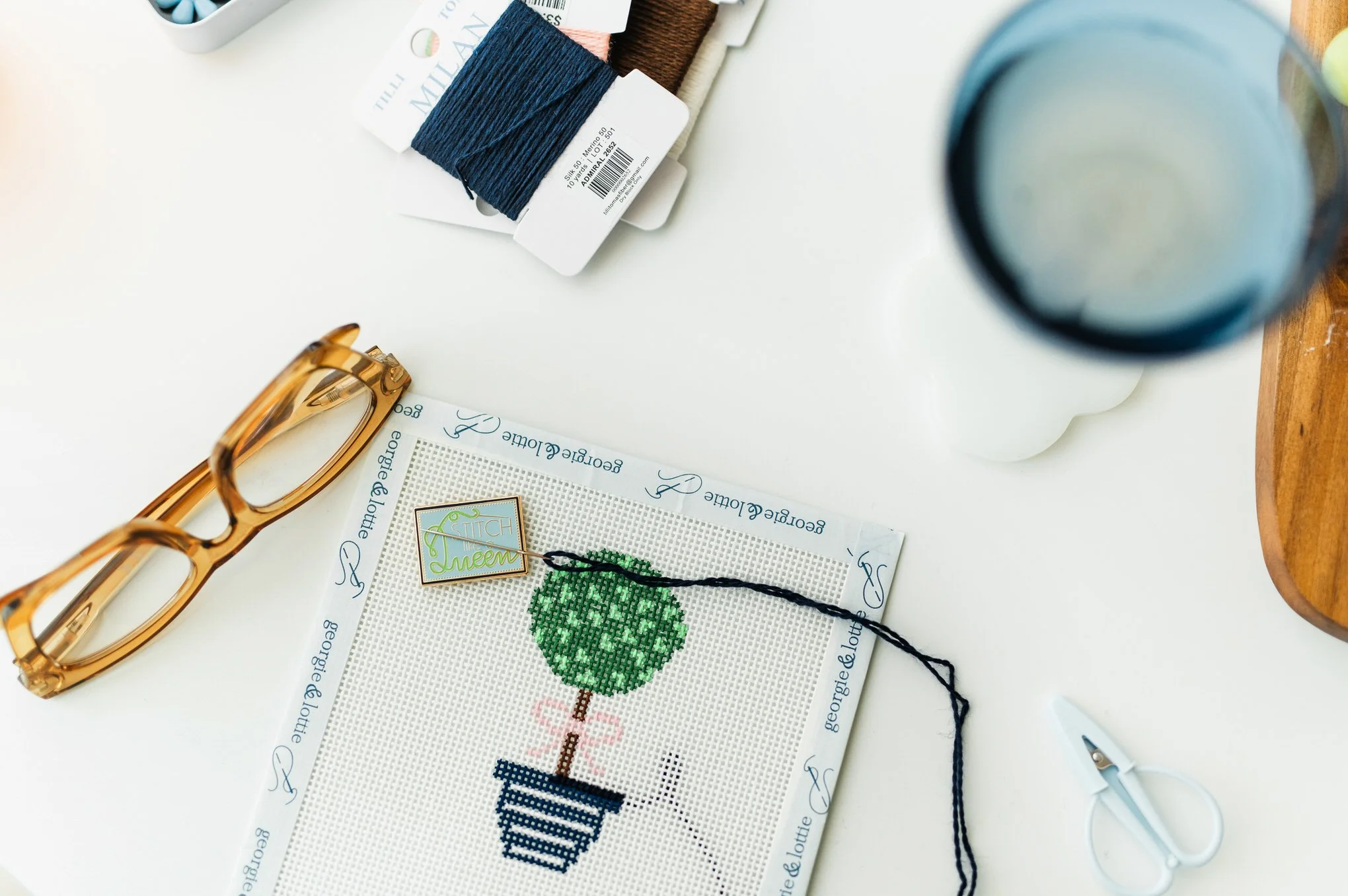 Cross-stitch embroidery project with a green heart design on white fabric, surrounded by sewing tools including scissors, thread, and fabric swatches, on a white surface.