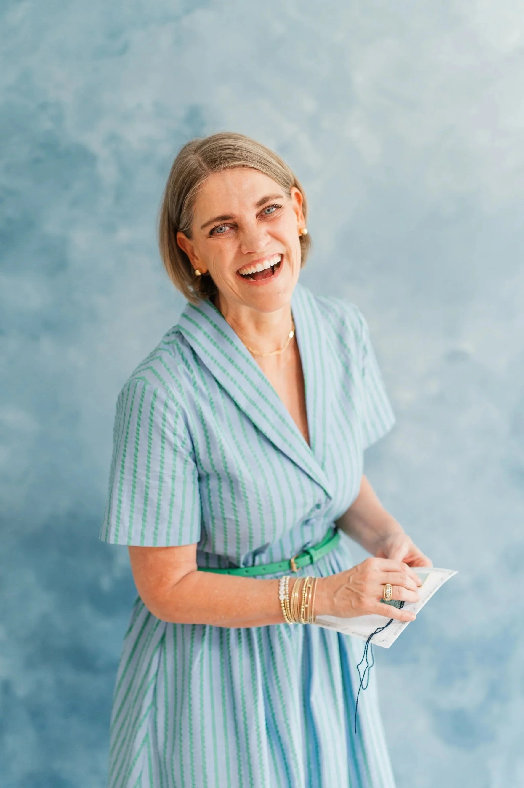 A smiling woman with short gray hair, wearing a green and white striped dress, gold jewelry, and holding a smartphone with earbuds against a blue textured background.