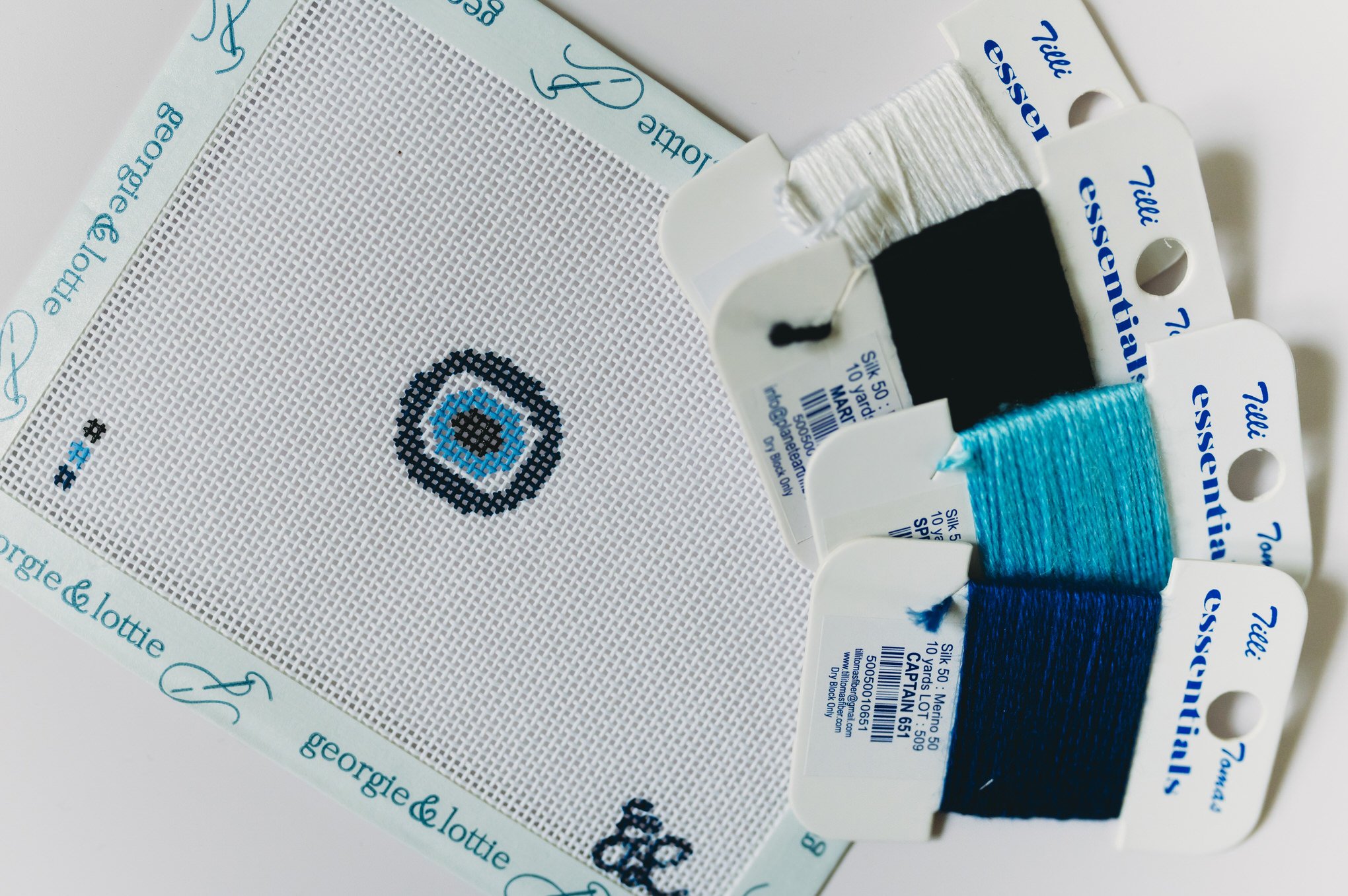 Hand-painted Georgie & Lottie evil eye mini needlepoint bag tag kit with blue concentric circle design and coordinating threads on 18 mesh canvas.