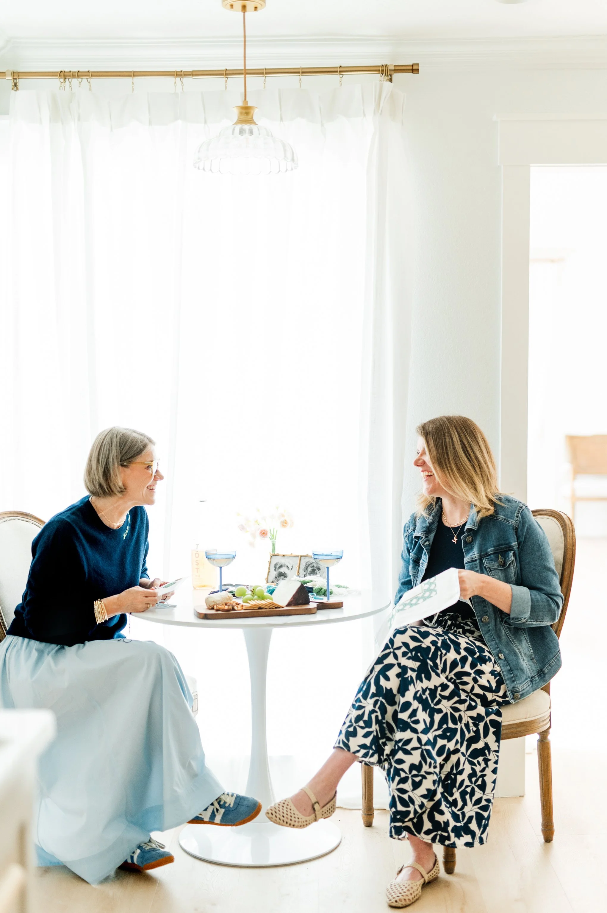 Two women laughing and talking at a round table with food and drinks during a daytime gathering in a bright, white room with white curtains and a hanging light fixture.