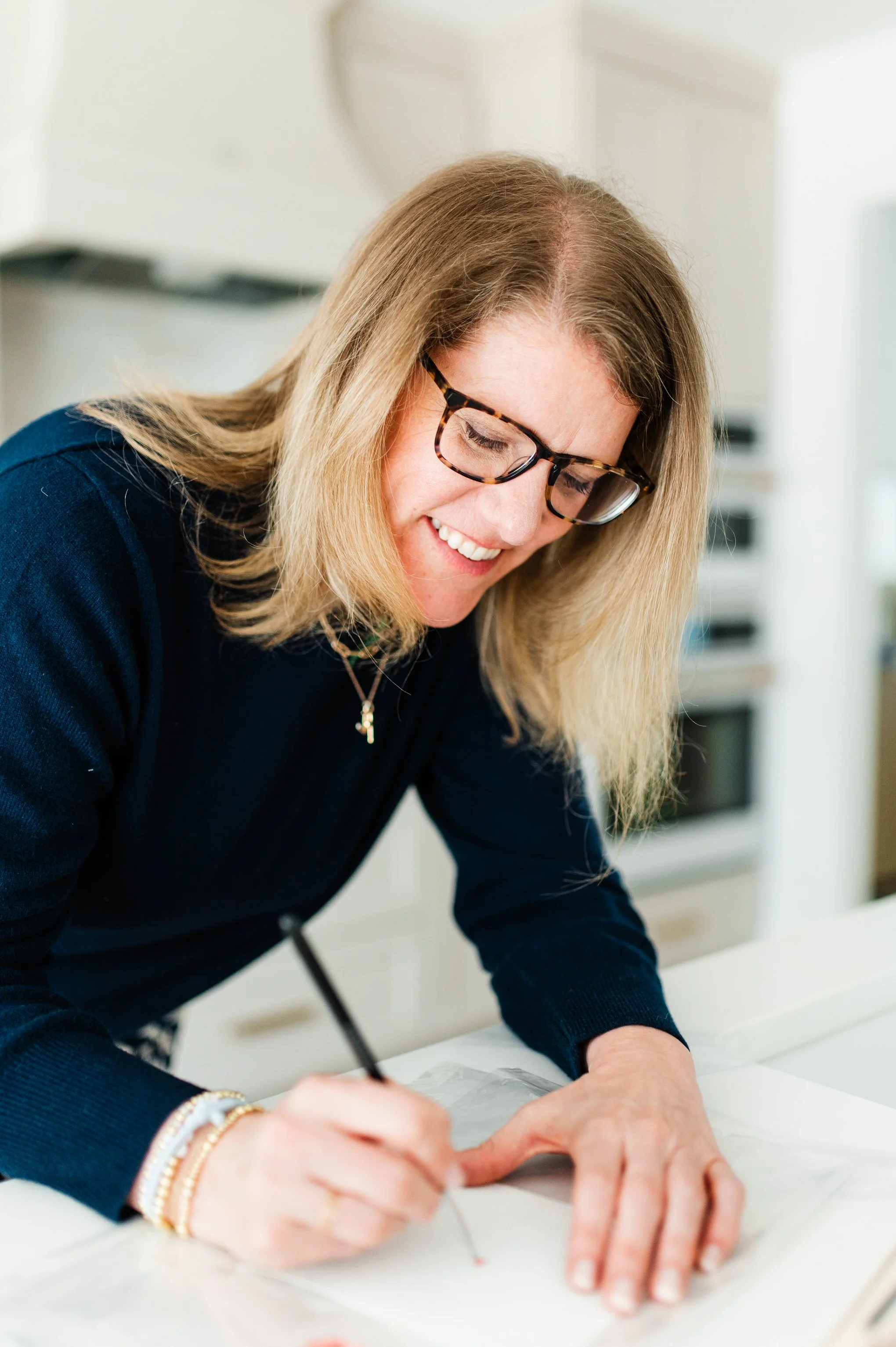 A woman with blonde hair, glasses, and wearing a navy blue top is smiling and writing or drawing on paper at a white table in a bright room.
