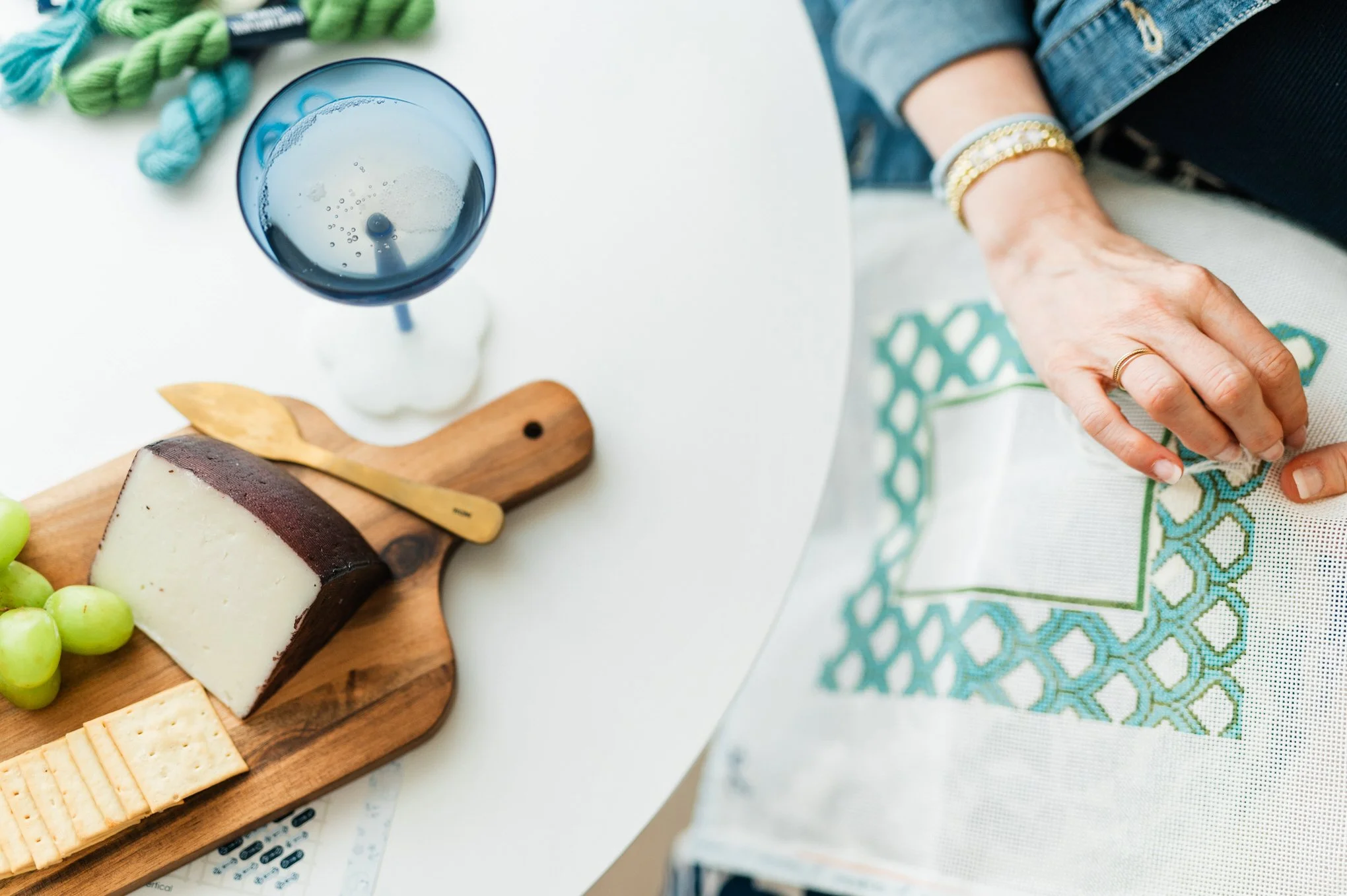 Top view of a blue wine glass with a beverage, a wooden cheese board with various cheeses, green grapes, and crackers, a hand resting on a table covered with a printed tablecloth, and a person's arm wearing a denim jacket and gold bracelet.