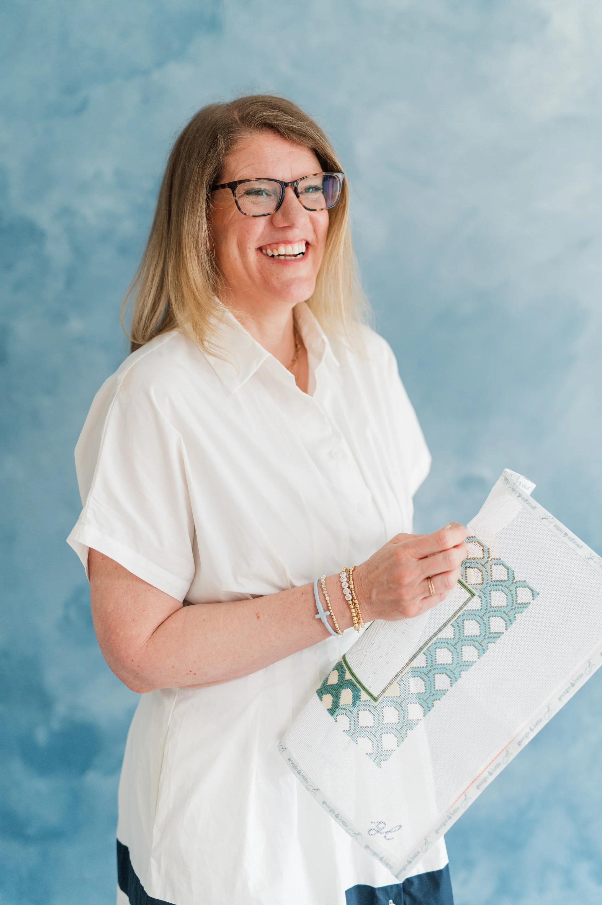 A woman with glasses and blonde hair, wearing a white shirt, holding a piece of cross-stitch embroidery pattern in front of a blue background.