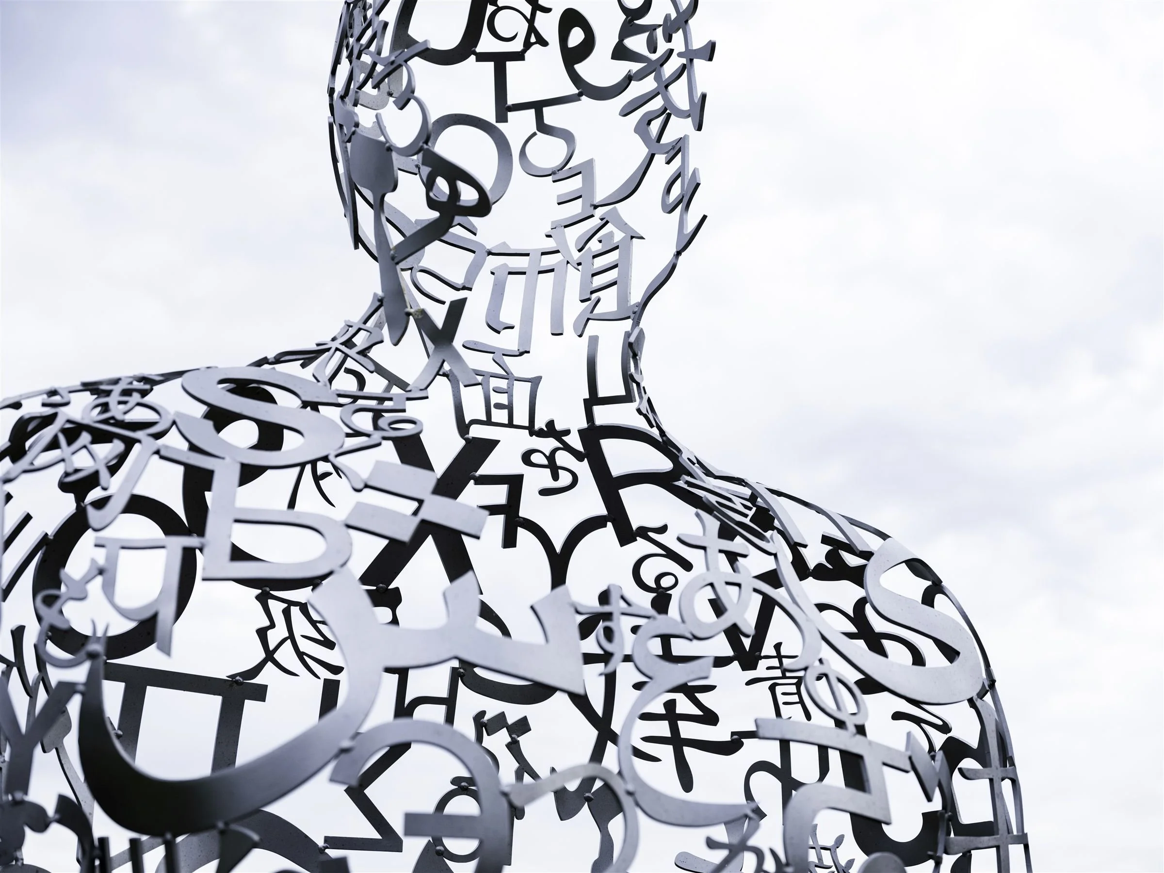 A sculpture of a human figure made from metal letters cutouts, seen from below against a cloudy sky.