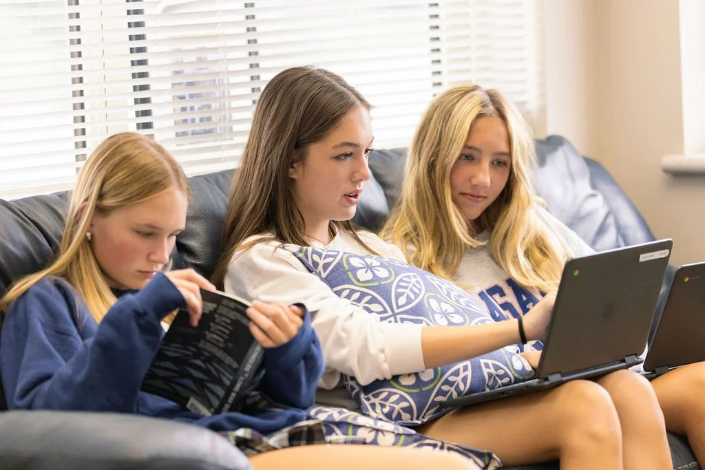 Three teenage girls are sitting on a couch, using laptops and reading a book in a living room with blinds on the window behind them.