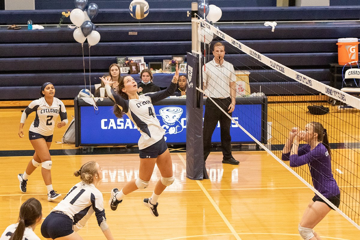 A volleyball game in progress with players from both teams near the net. One player is jumping to hit the ball, while the opponent crouches to receive the serve. The gym has blue bleachers and balloons.