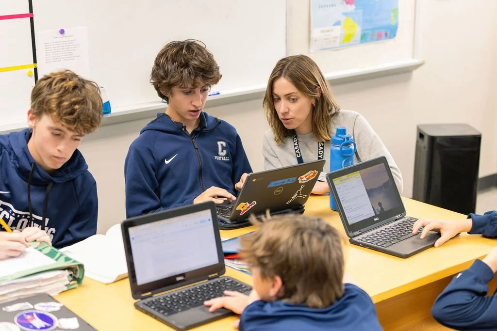 A classroom with students working on laptops at a table, with a teacher assisting a student.