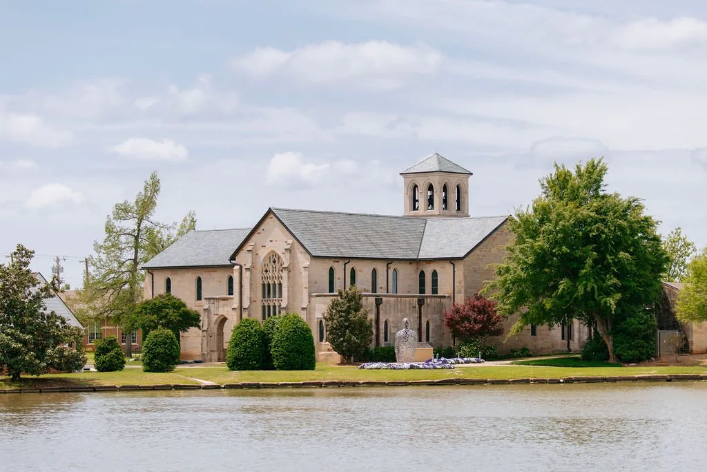 A historic church with a large stone facade, gothic windows, and a tower, situated along a body of water surrounded by trees and greenery.