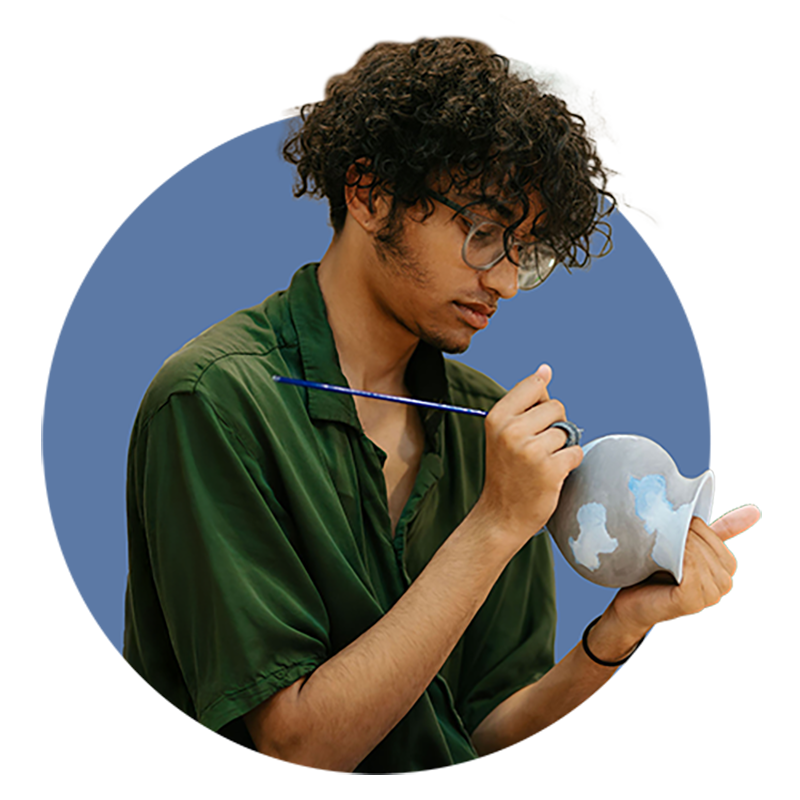 A person with curly hair and glasses painting a cloud design on a white ceramic vase.
