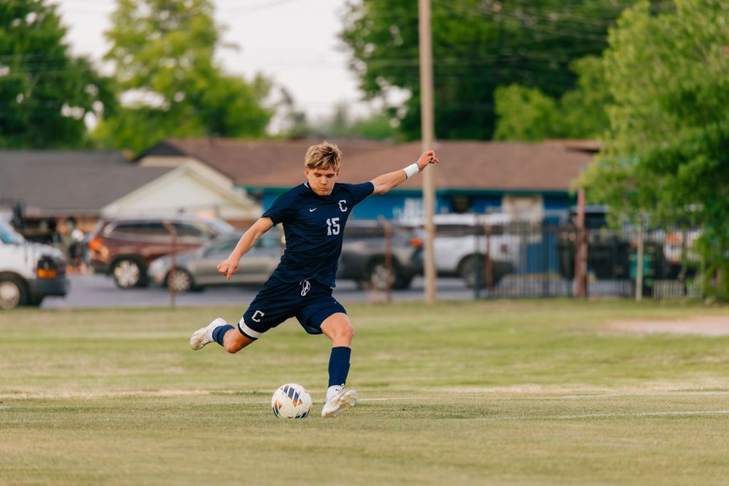 A young soccer player in a navy uniform kicking a soccer ball on a grassy field with trees and houses in the background.
