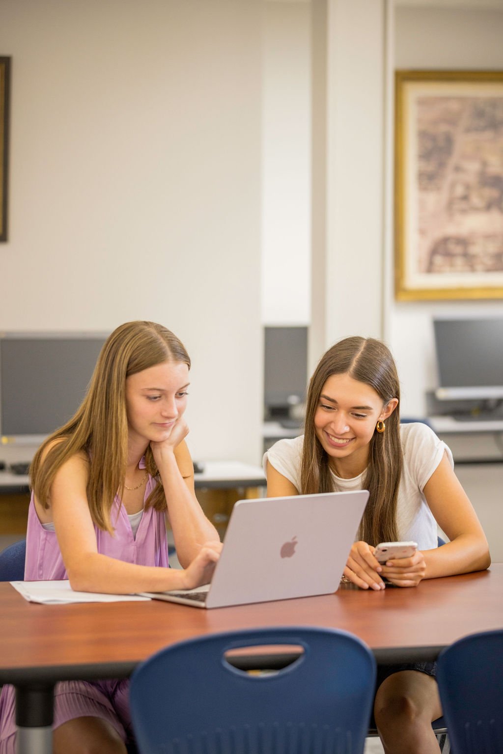 Two young women sitting together at a table in a classroom, looking at a laptop and a phone and smiling.