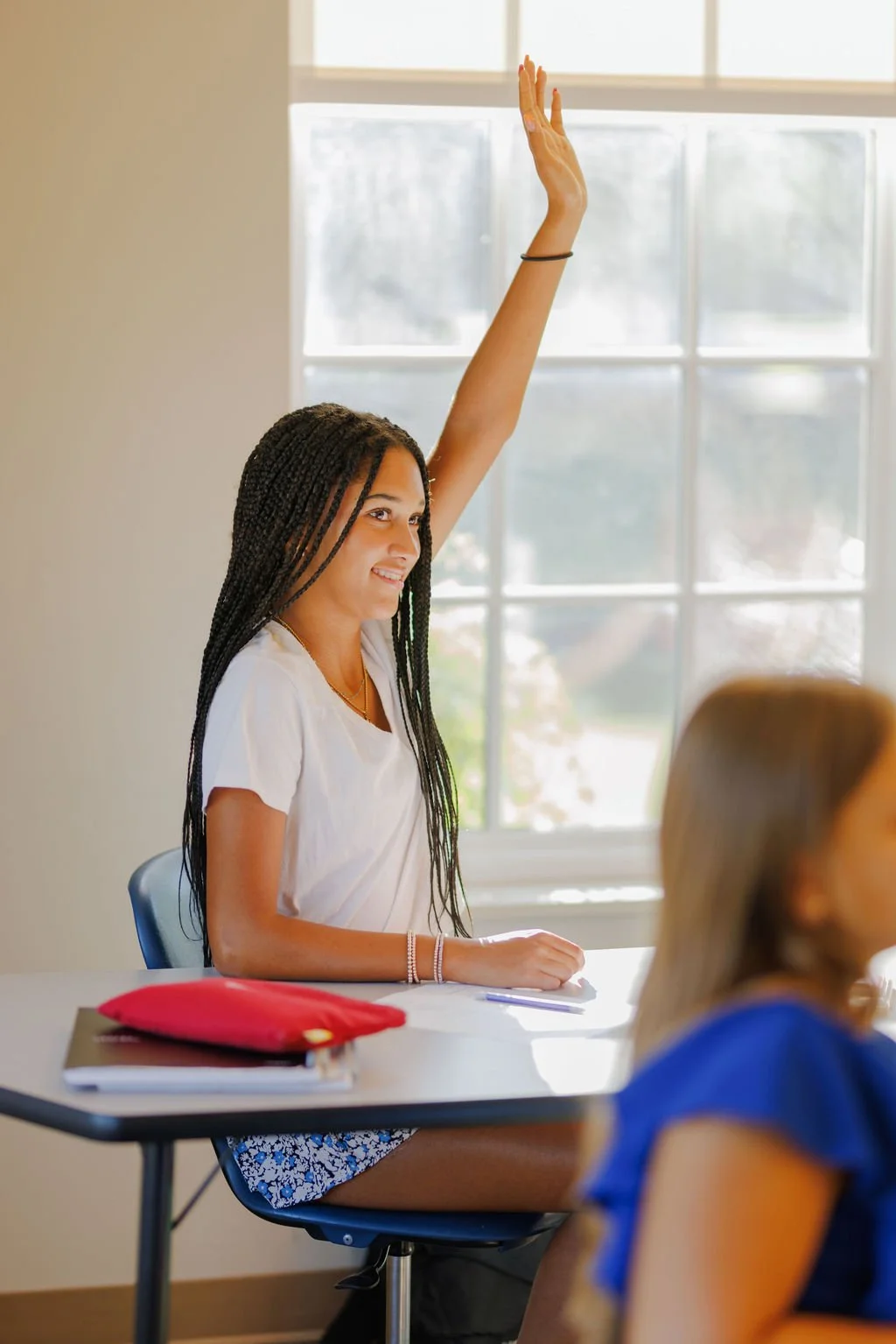 A girl with long braids, wearing a white t-shirt and floral shorts, raising her hand in a classroom during daytime, with sunlight coming through large window in the background.