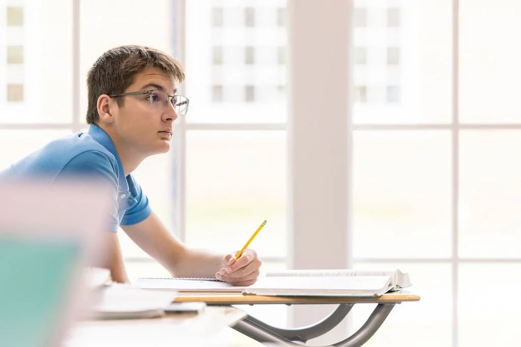 A young man with glasses sitting at a school desk, holding a pencil, looking thoughtfully out the window in a bright classroom.