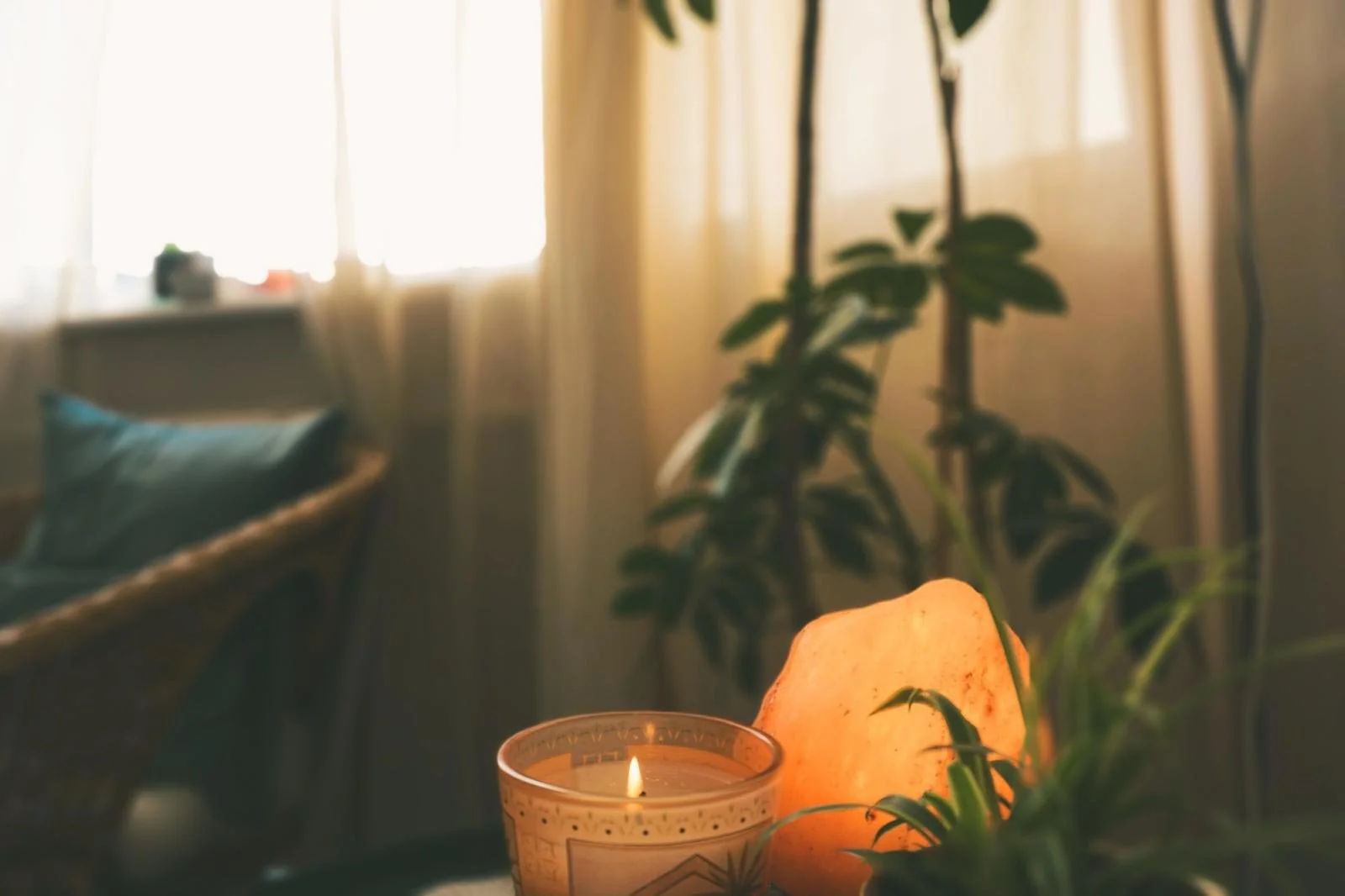 A cozy interior corner with a lit candle and a Himalayan salt lamp on a table, with potted plants and curtains in the background.
