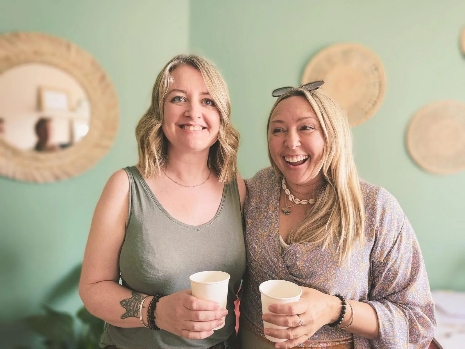 Two women smiling and holding cups, standing close together indoors with decorative mirrors on a green wall.