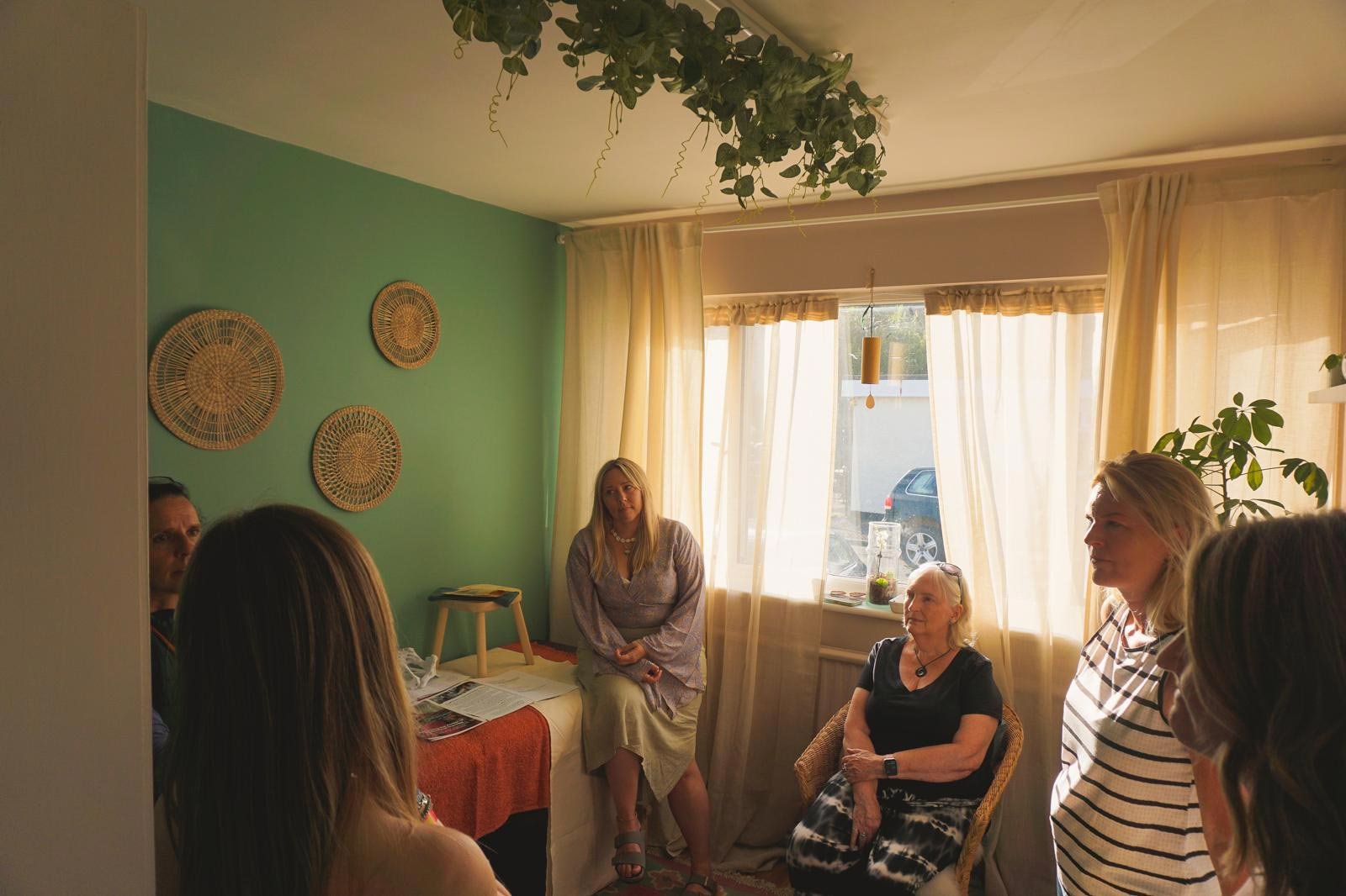 Six women seated and standing in a cozy living room with green wall, cream curtains, and decorative wicker wall art, engaged in conversation during daytime.