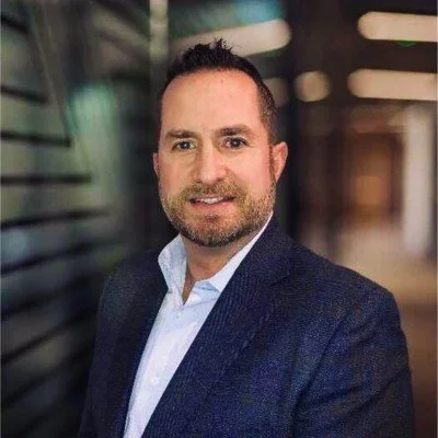 Portrait of a man with dark hair, beard, wearing a dark blazer and white shirt, standing in a modern building with glass and metal accents.