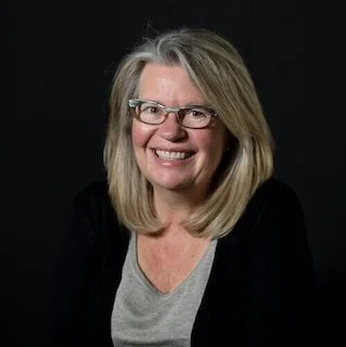 Lisa Grize smiling, and dressed in a gray top with a black jacket against a dark background.