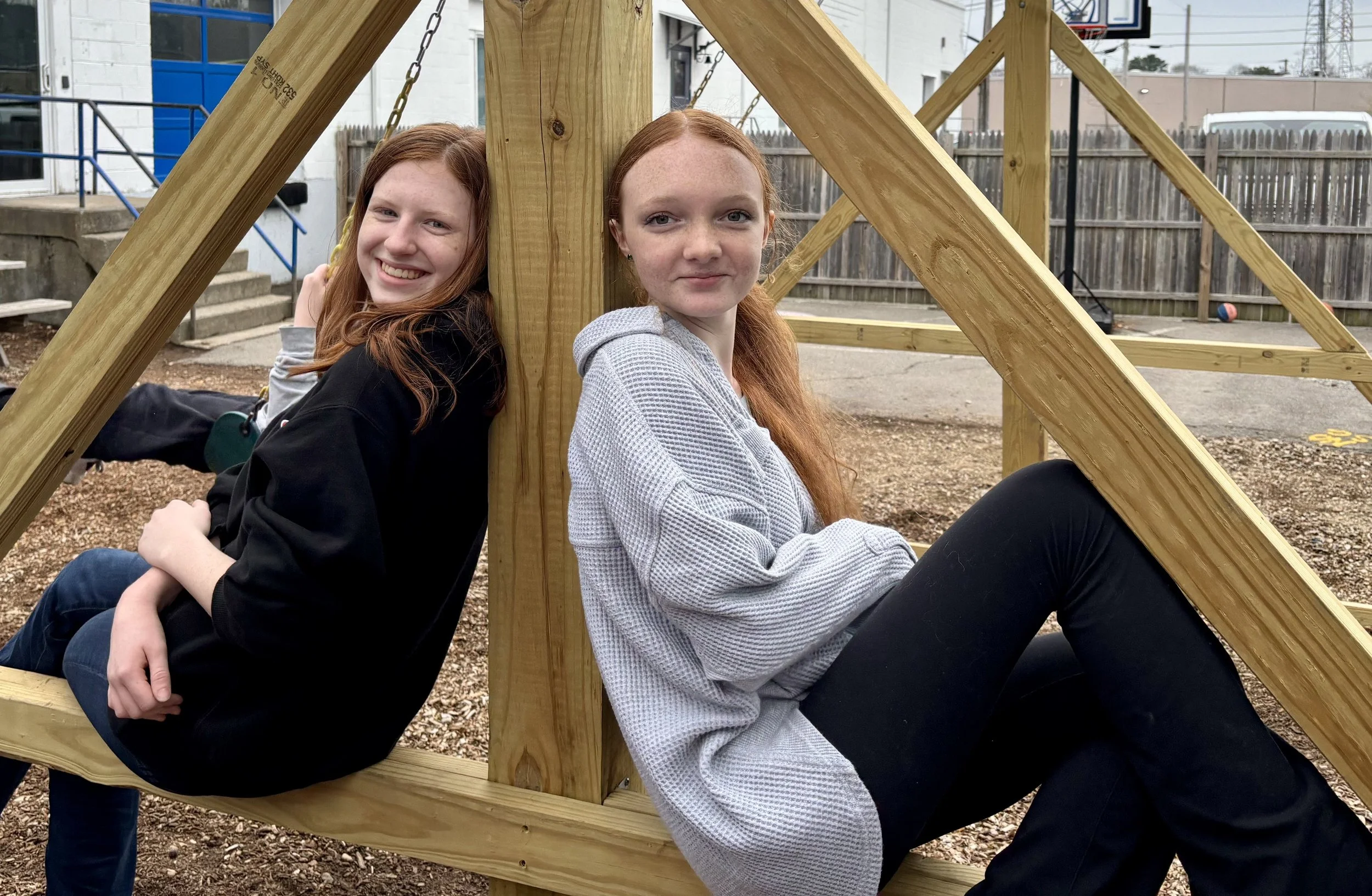 Two young women with red hair sitting on a wooden playground structure, smiling at the camera.