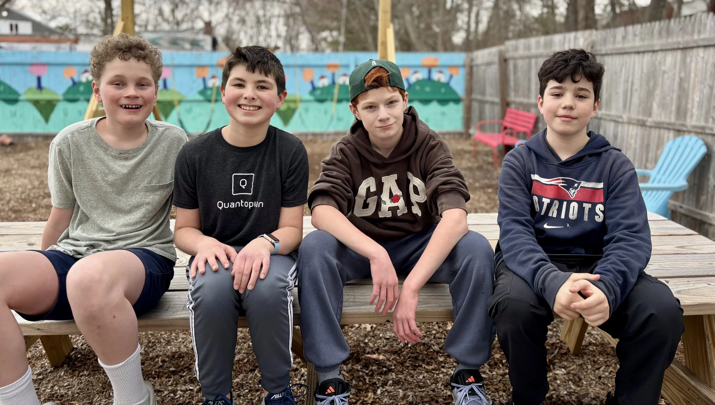 Four boys sitting on a wooden bench in an outdoor playground, with a colorful mural and chairs in the background.