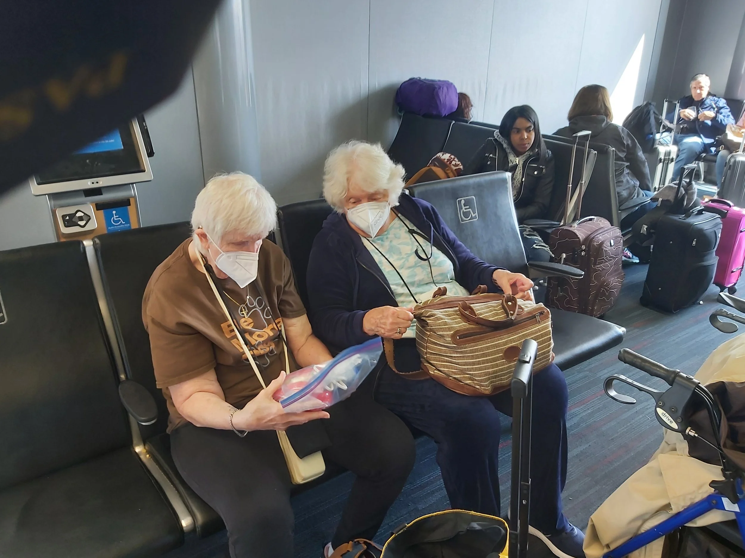 Two women with white hair sitting on black airport chairs with a wheelchair accessibility symbol visible on a chair and a walker in the foreground.