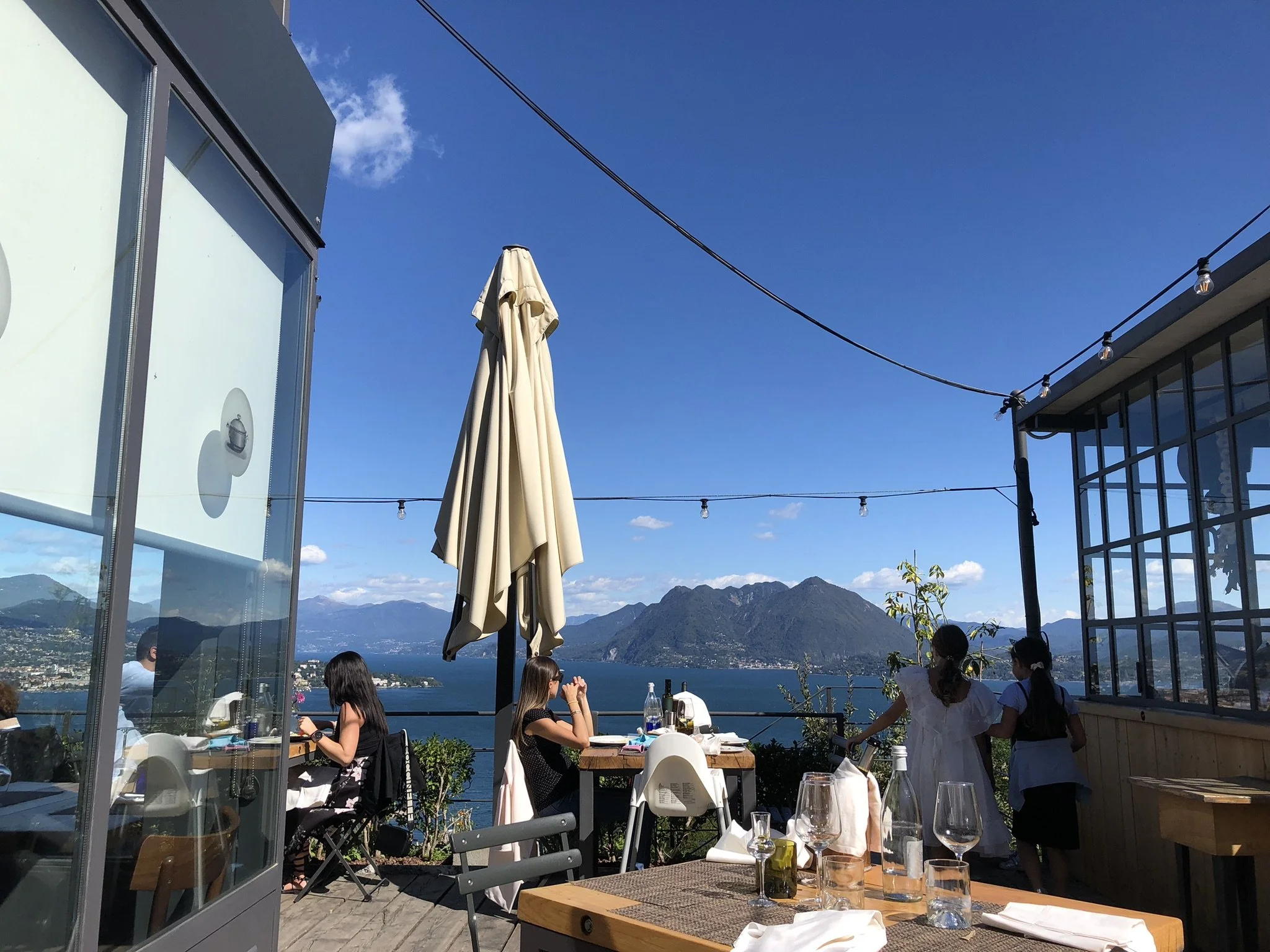Wooden deck and wooden restaurant tables with a collapsed umbrella with the blue water of Lake Maggiore in the background, with mountains and a deep blue sky with puffy white clouds at La Rampolina restaurant.