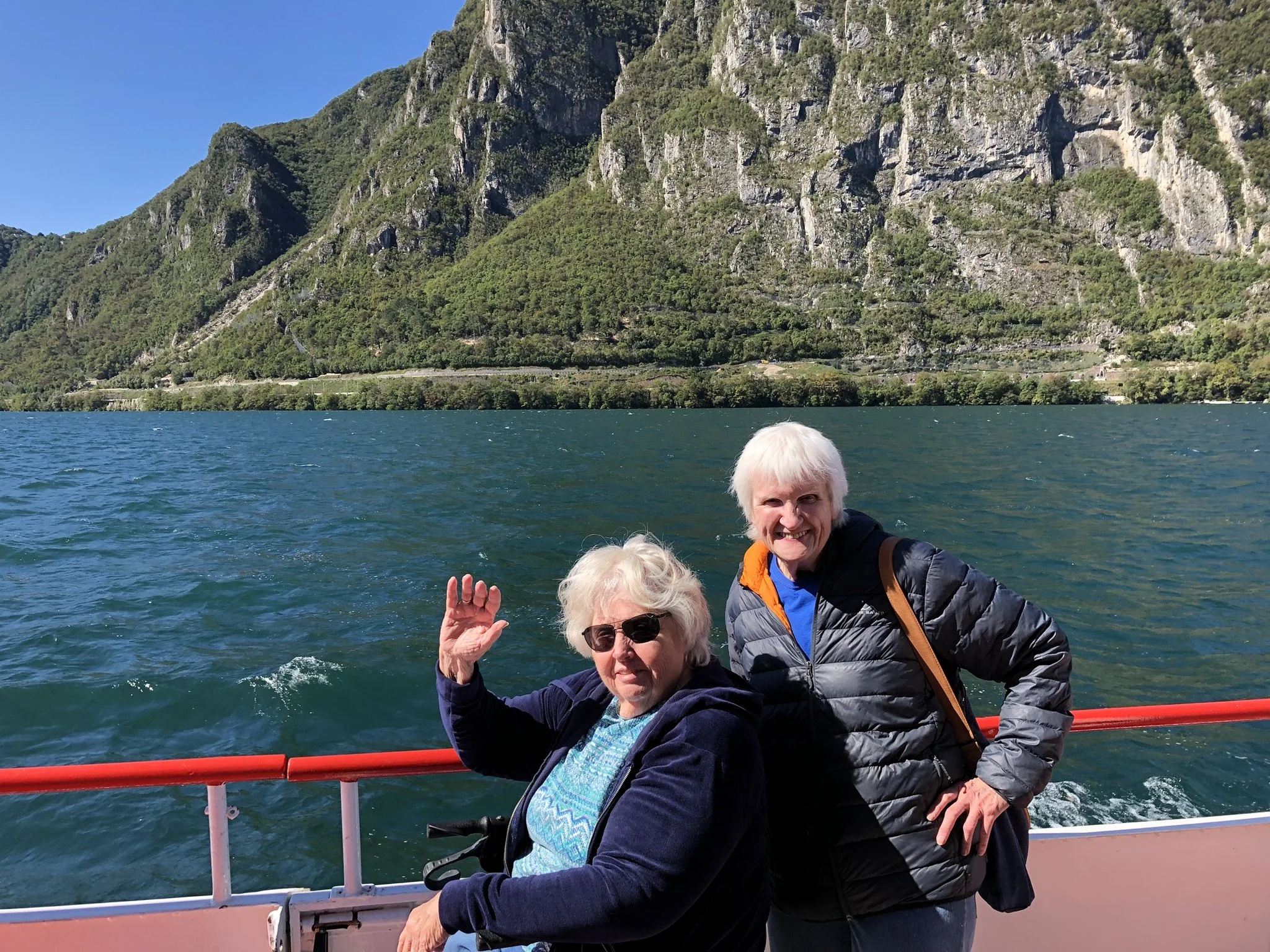 Two women with white hair, one with dark sunglasses, in front of a red ferry railing, blue green water with white caps of Lake Lugano in the background, with steep hills with rocks and greenery in the far background.