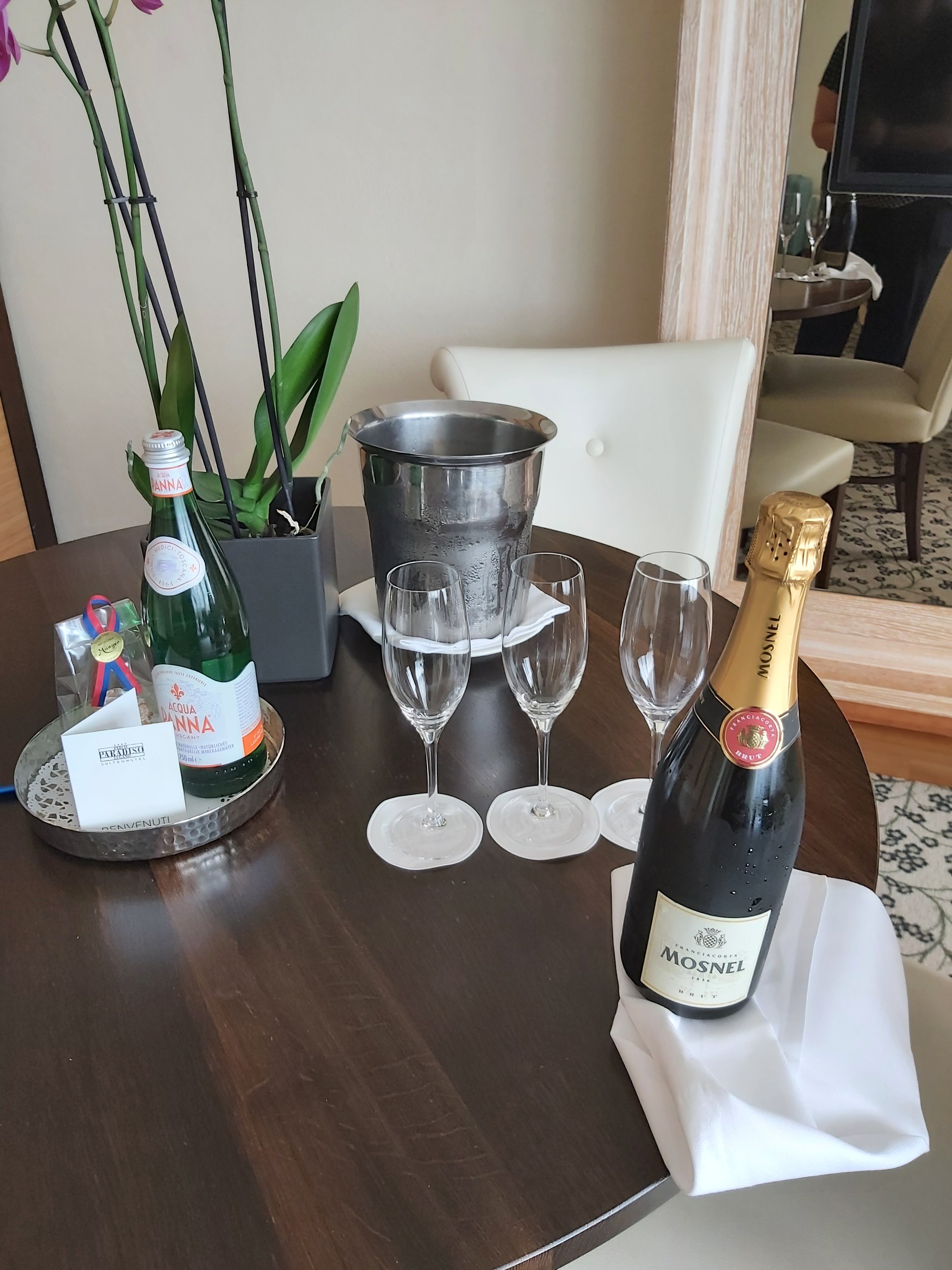 Dark wood table with silver ice bucket, gold foil topped bottle of wine, water bottle, three champagne flutes, and the base of an orchid visibile.