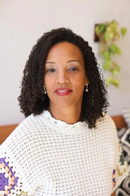 A woman with curly black hair wearing a white crocheted sweater with purple and yellow details, sitting in a room with a white wall and a hanging plant in the background.