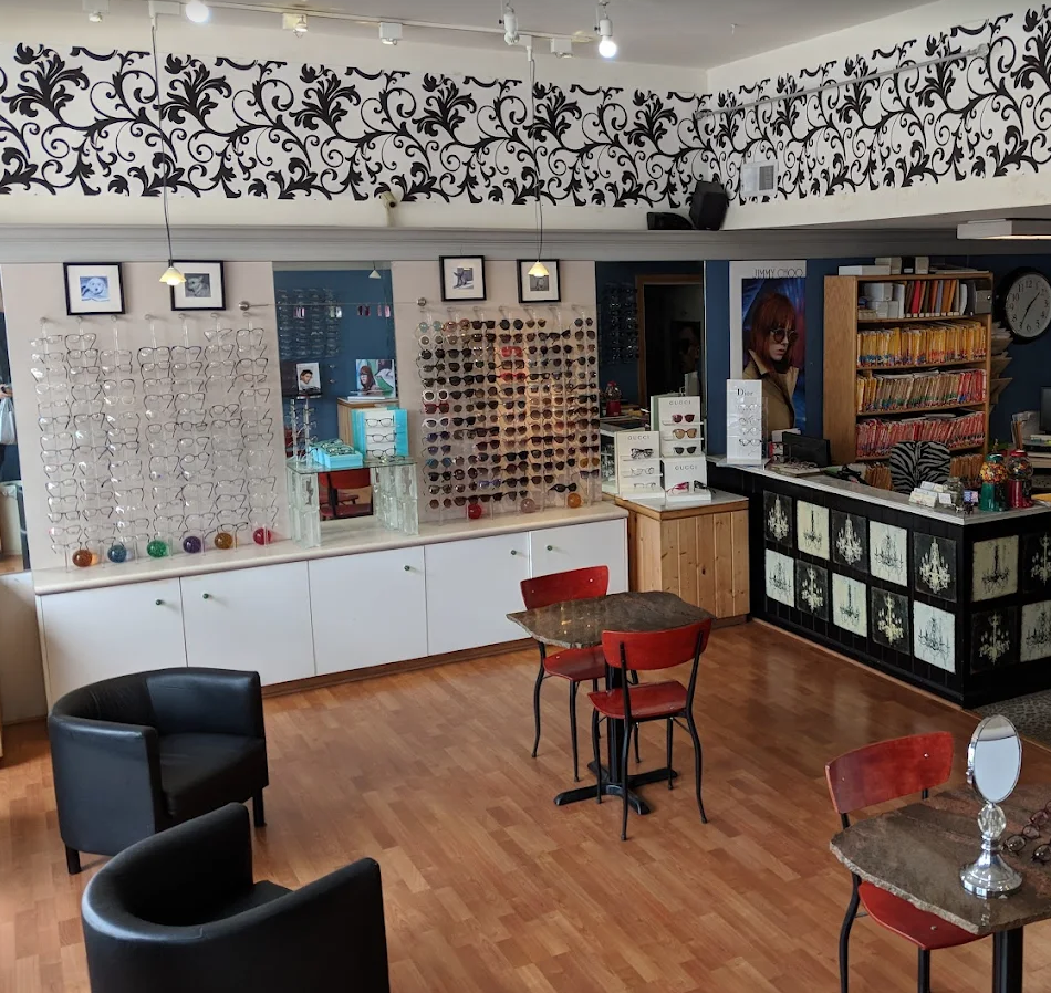Interior of an eyewear store with eyeglasses on display, a checkout counter with books and eyewear accessories, and small tables with red chairs, wooden flooring, and decorative black floral patterns on the upper wall.