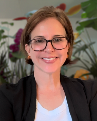 A woman with short brown hair, glasses, and a smile standing in front of a colorful mural with plants.