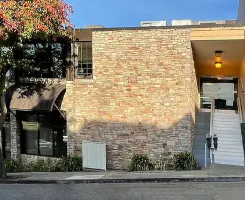 Exterior view of a modern building with brick and stone walls, large glass window, staircase, and a small landscaped area with plants, under a partly cloudy sky.