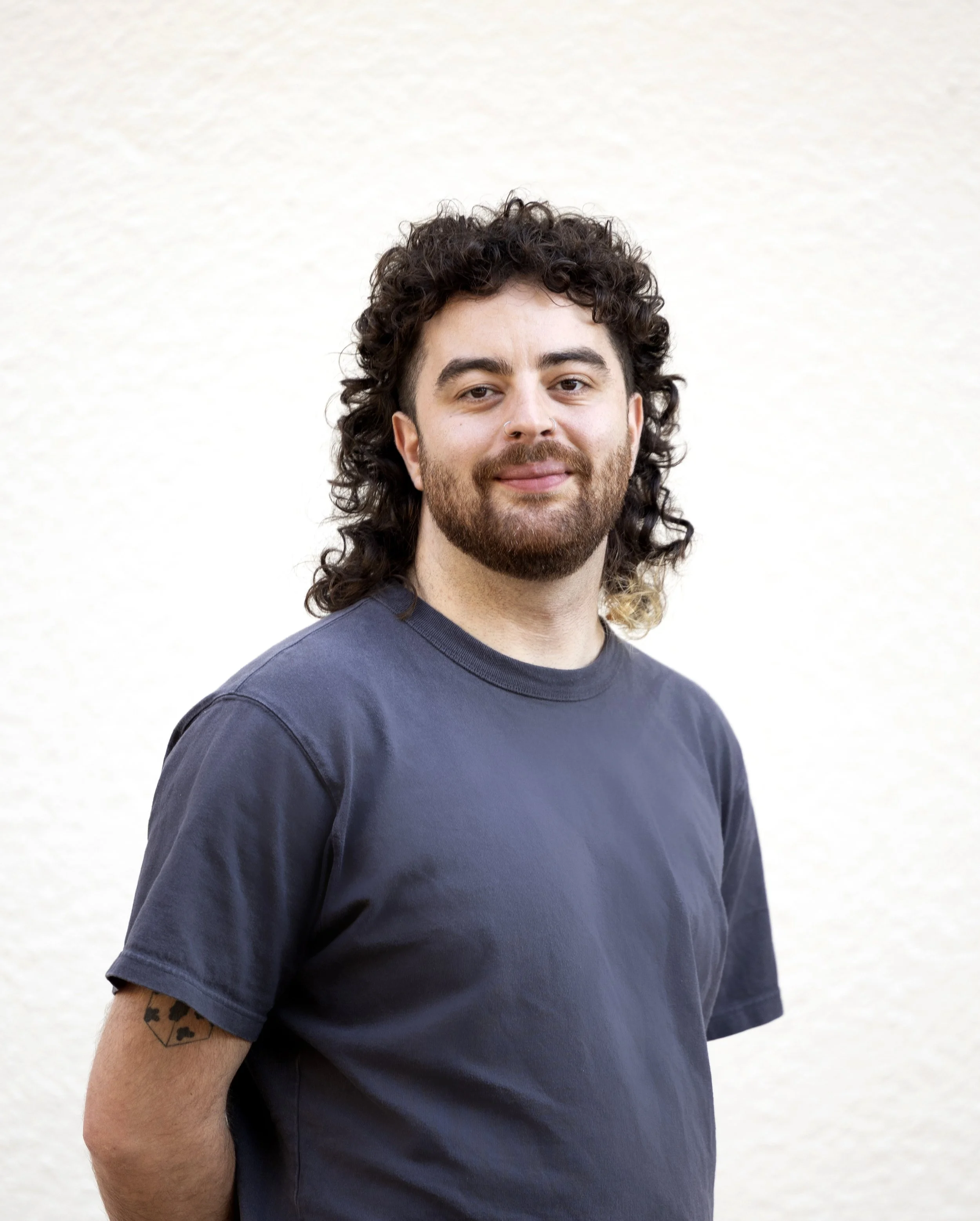 A man with curly hair, a beard, and a nose ring, wearing a dark t-shirt, standing outdoors against a plain background.