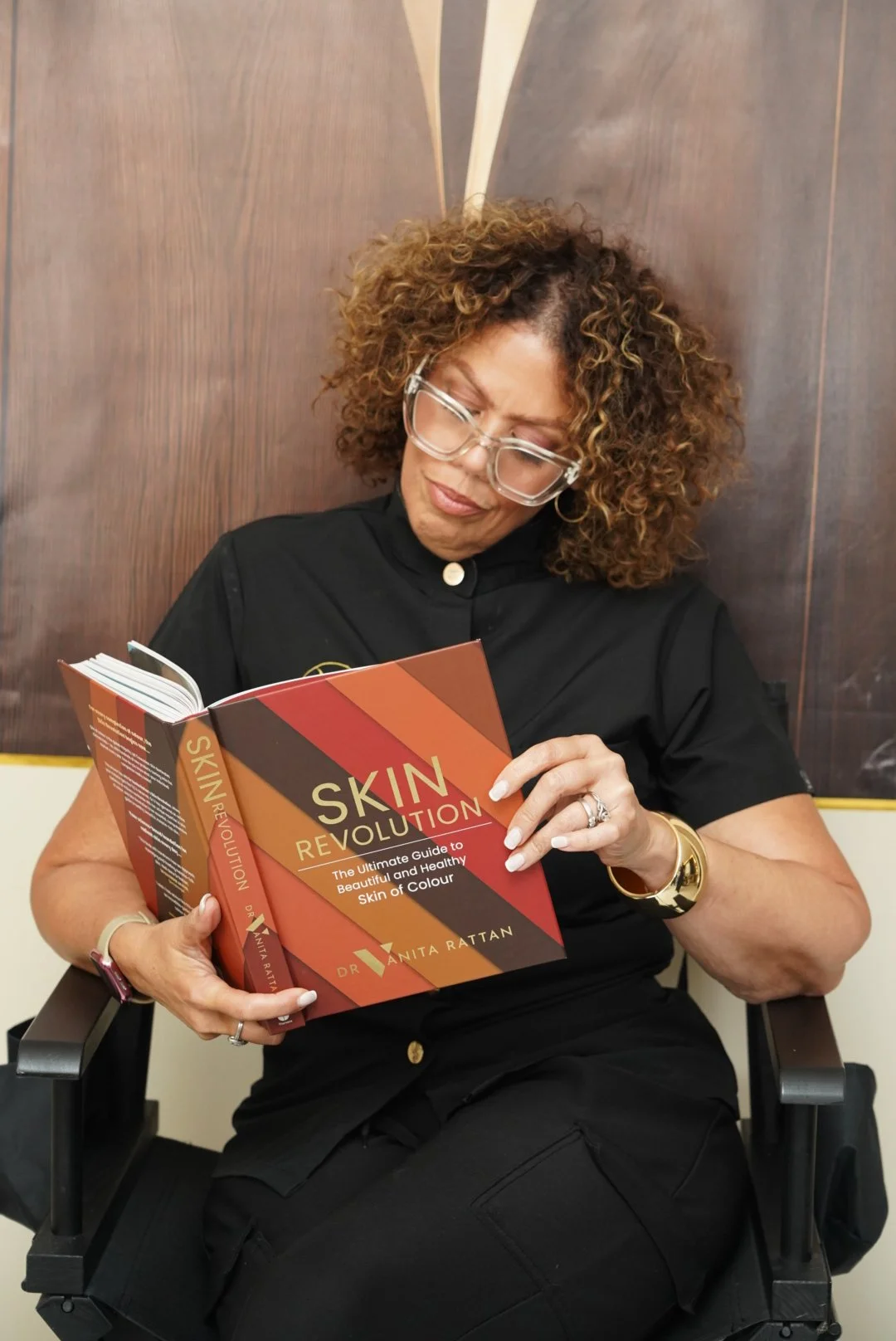 Woman with curly hair and glasses sitting in a wheelchair, reading a book titled 'Skin Revolution' by Dr. Anita Rattan, in front of a wooden wall.
