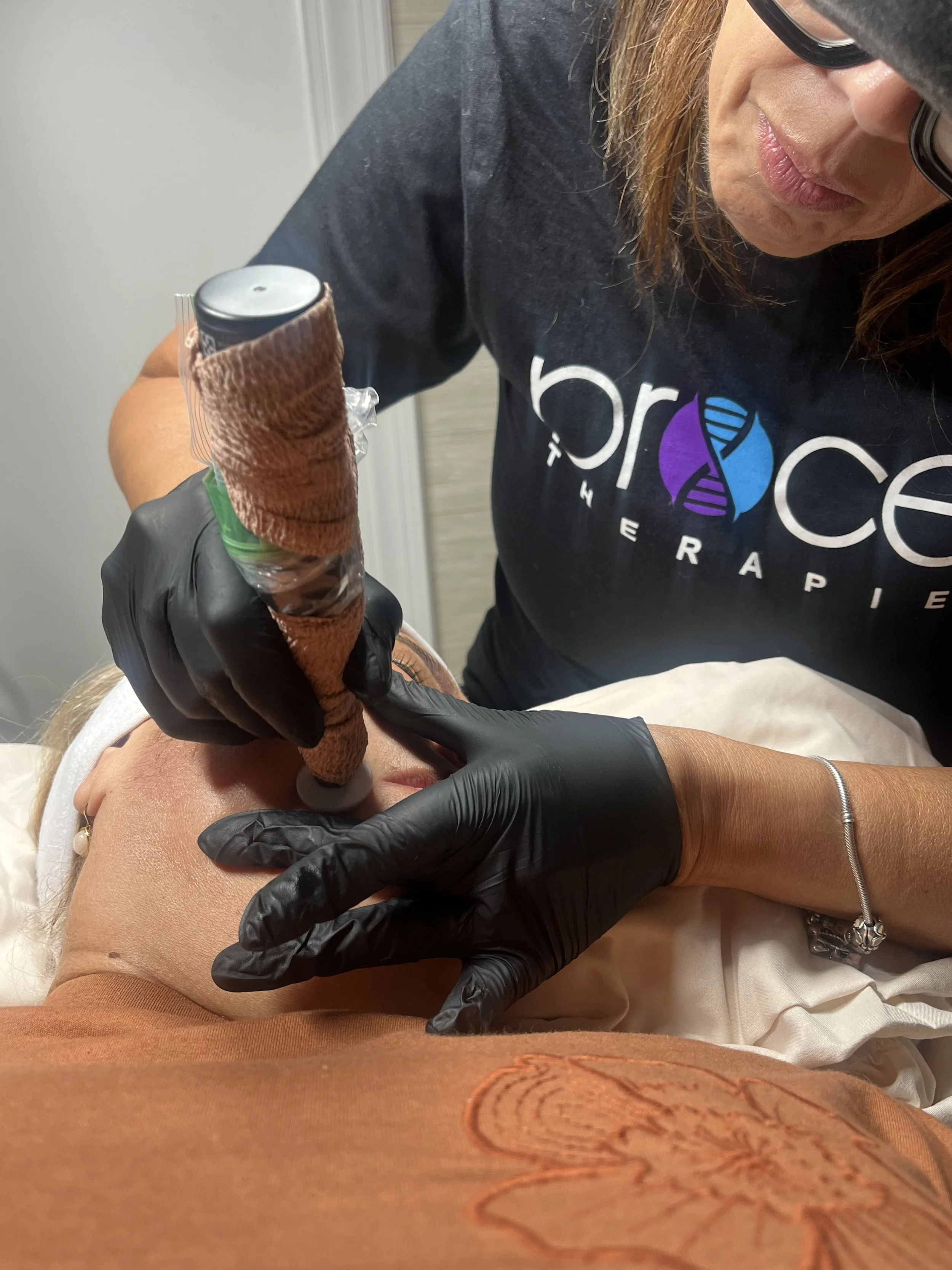 A healthcare professional wearing black gloves and a dark shirt with a logo labeled "Progress Therapeutic" is administering a cosmetic or medical injection into a patient’s arm. The patient is lying down, wearing a white shirt and a bracelet on their wrist.