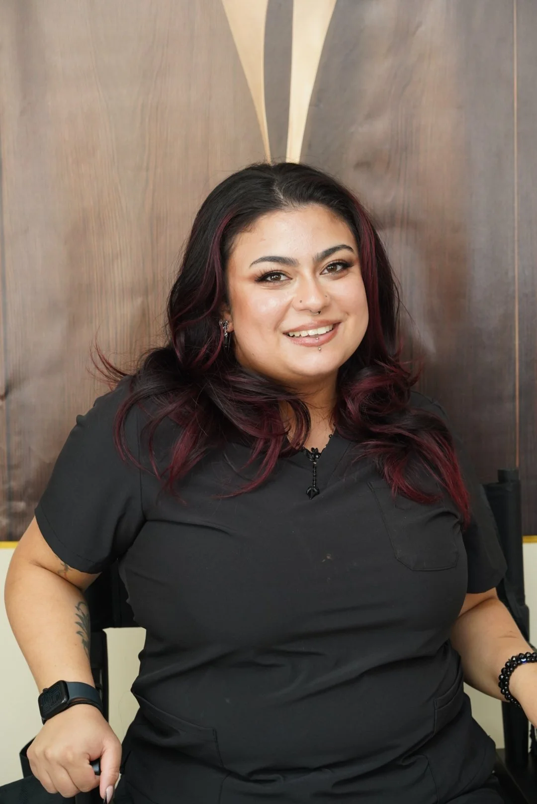 A woman with dark hair with red highlights, smiling, wearing black scrubs, sitting against a wooden background.