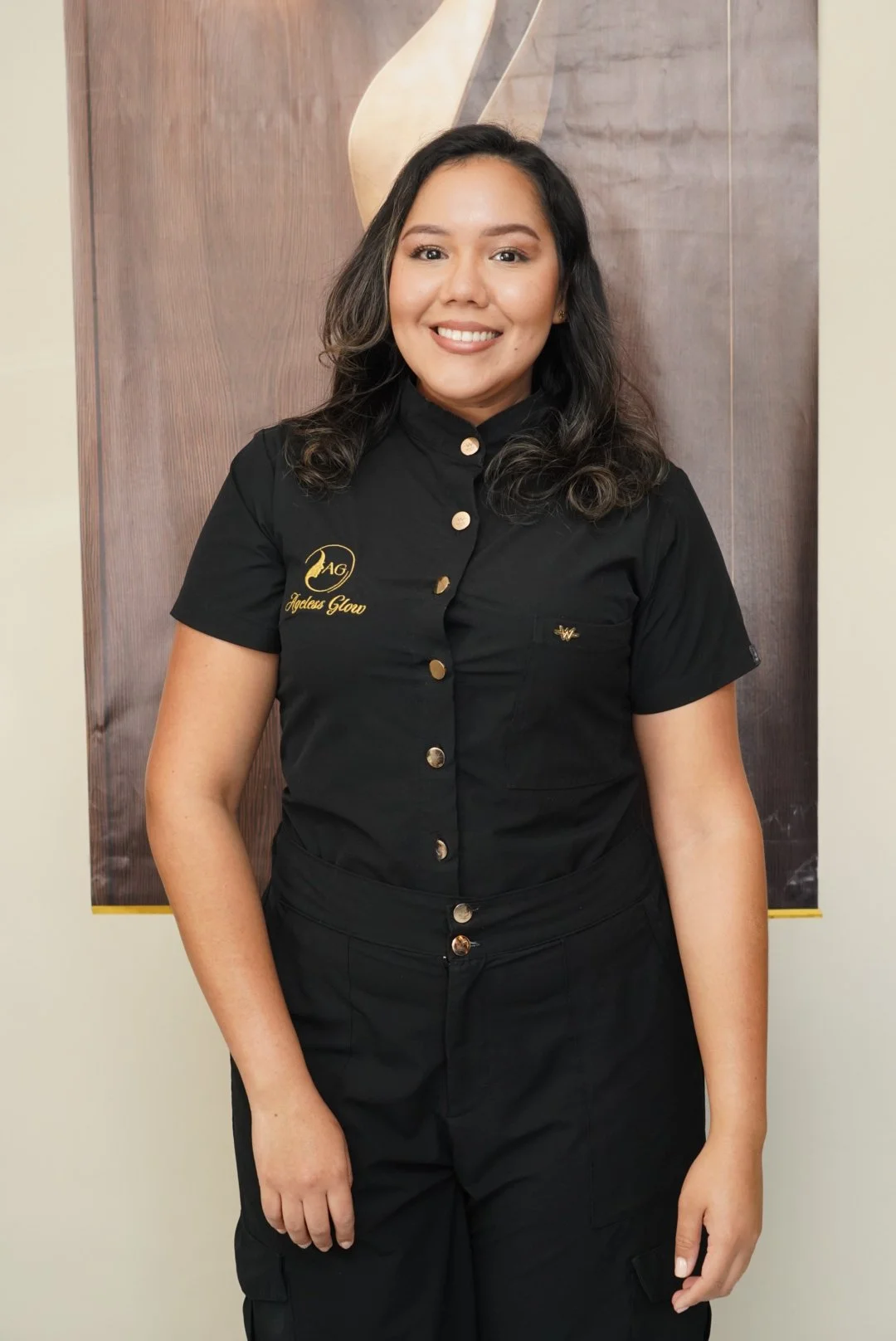 A smiling woman with dark, wavy hair wearing a black uniform shirt with gold buttons and embroidery that reads "Ageless Glow" and stands in front of a wooden panel with a decorative element.