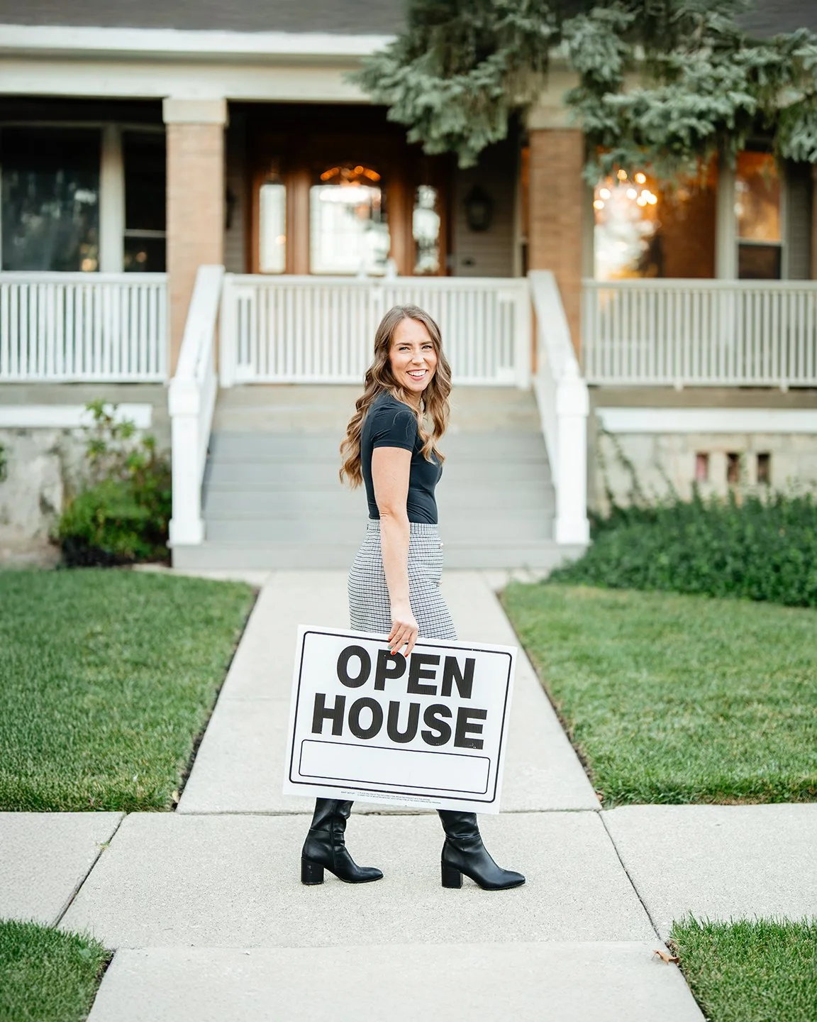 Young woman with blonde hair lying on a tan leather sofa with her legs extended and wearing chunky black shoes, a green checkered dress, a white long-sleeve shirt, and white socks, in a bright, minimalistic room with a green plant.