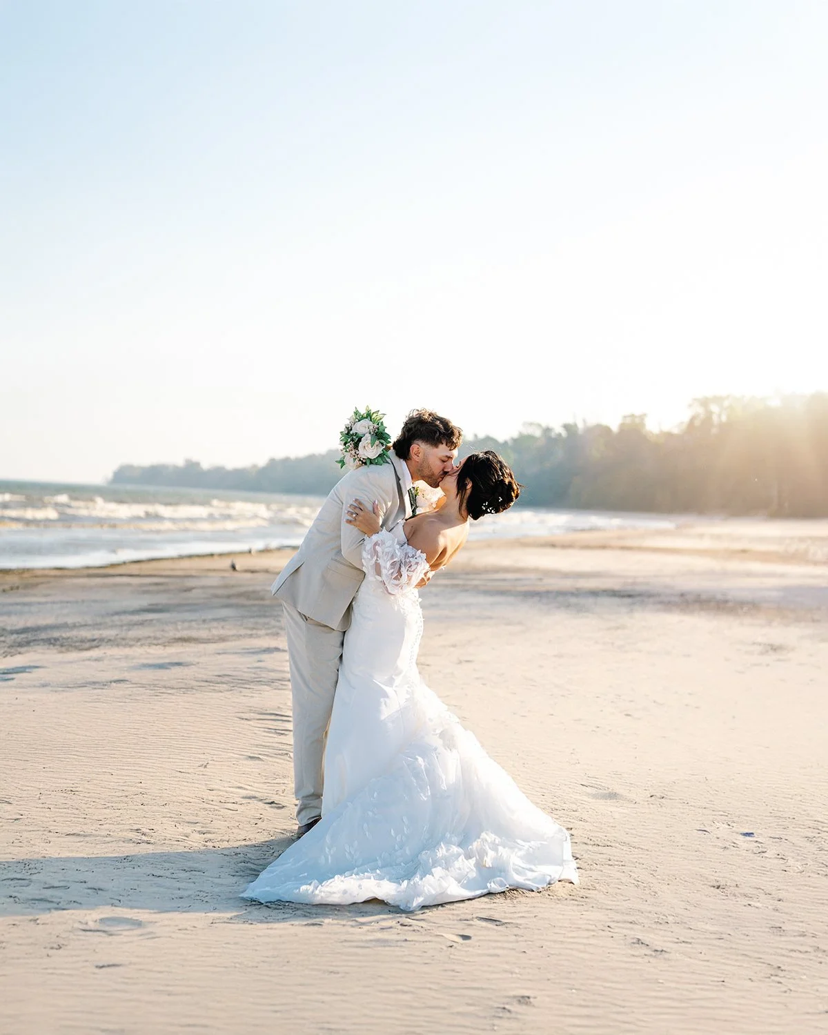 A bride and groom stand close to each other next to a vintage white car in a field of yellow flowers. The bride wears a white lace wedding gown, holding a bouquet, and leans towards the groom, who is dressed in a gray suit. They are about to kiss under a cloudy sky.
